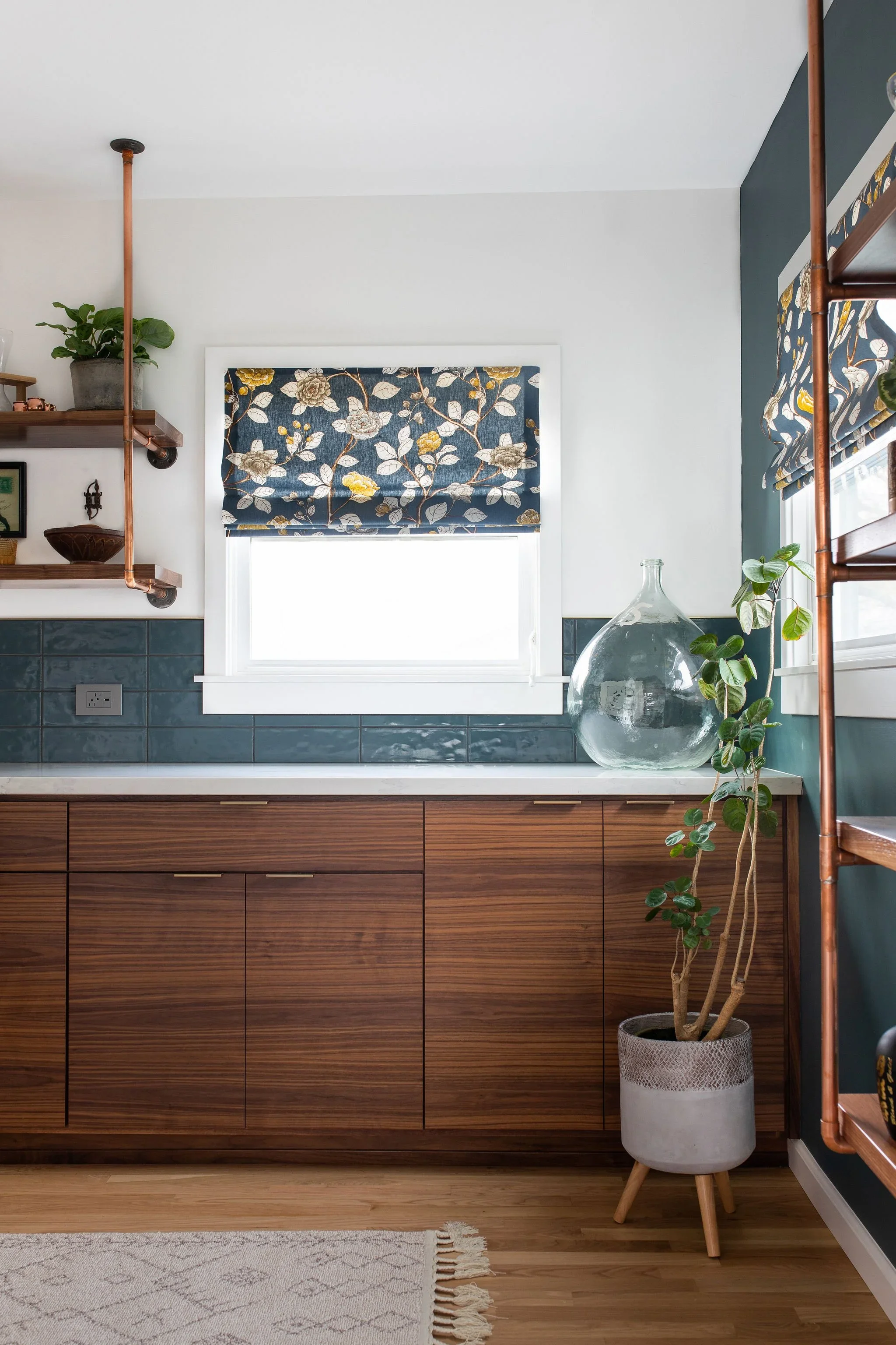 A kitchen with dark teal tiled backsplash, wooden cabinetry, a window with a floral Roman shade, and decorative items including plants, shelves, and a large glass pitcher.