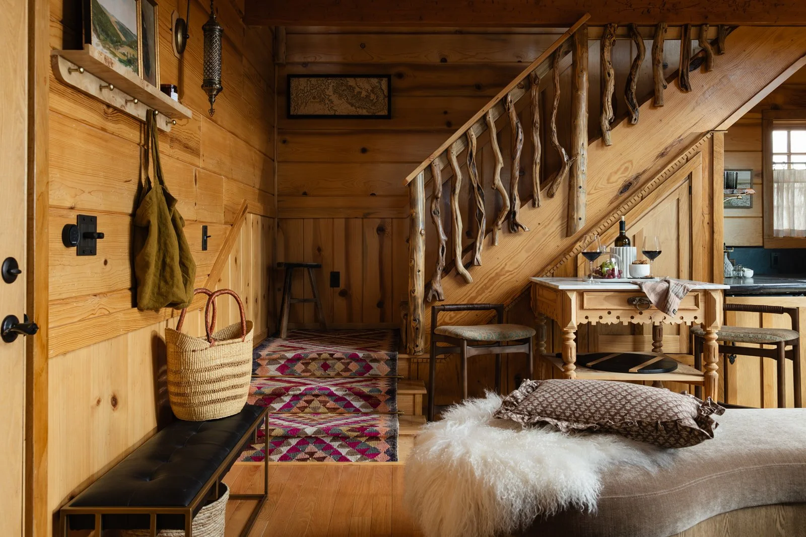 Interior of a cozy rustic cabin with wooden walls and furnishings, a staircase with a branch-inspired railing, a small table with wine and food, and a bed with a pillow and fur throw.
