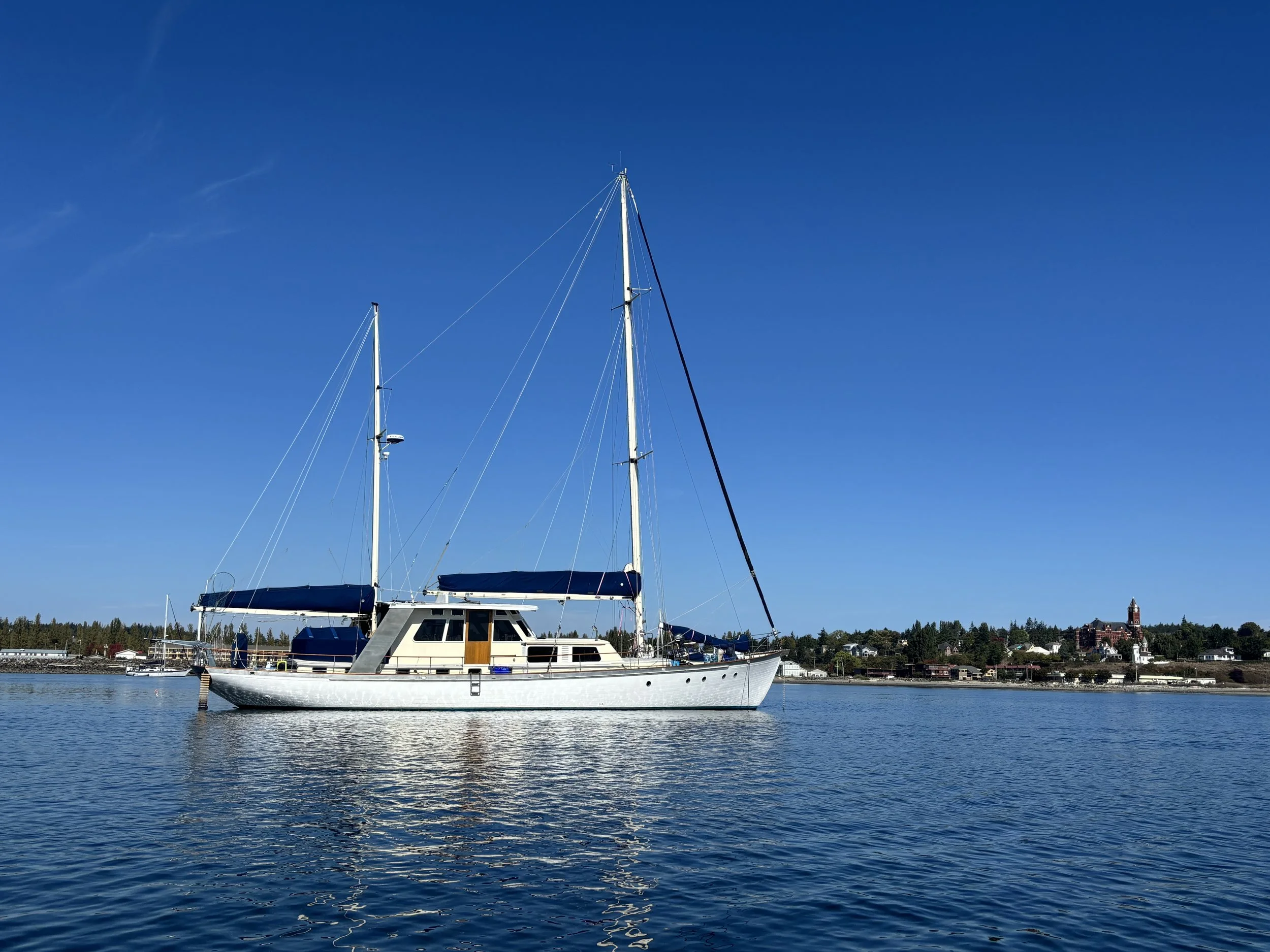 Sailboat floating on calm water with a shoreline and buildings in the background under a clear blue sky.