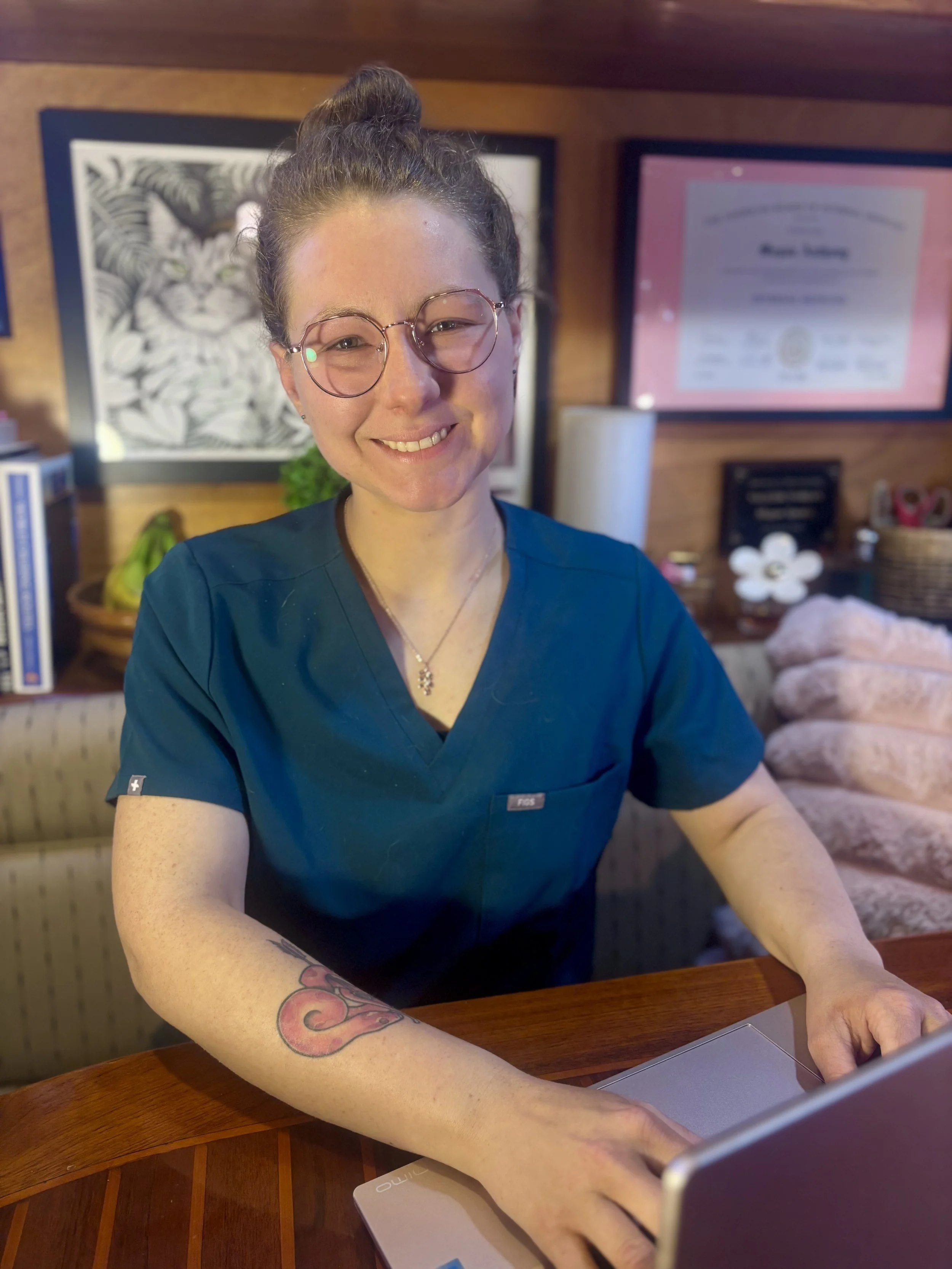 Portrait photo of Dr. Megan Anthony DO at her home office. A caucasian woman wearing a set of teal scrubs with rose gold glasses and light brown hair tied into a bun. She is smiling while working on a laptop at a wooden table.