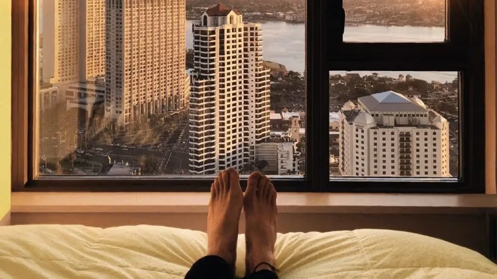 View from a high-rise window showing tall buildings, a river, and a cityscape at sunset, with a person's feet visible in the foreground on a bed.