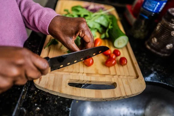 Person chopping cherry tomatoes on a wooden cutting board with lettuce and cucumber in the background.