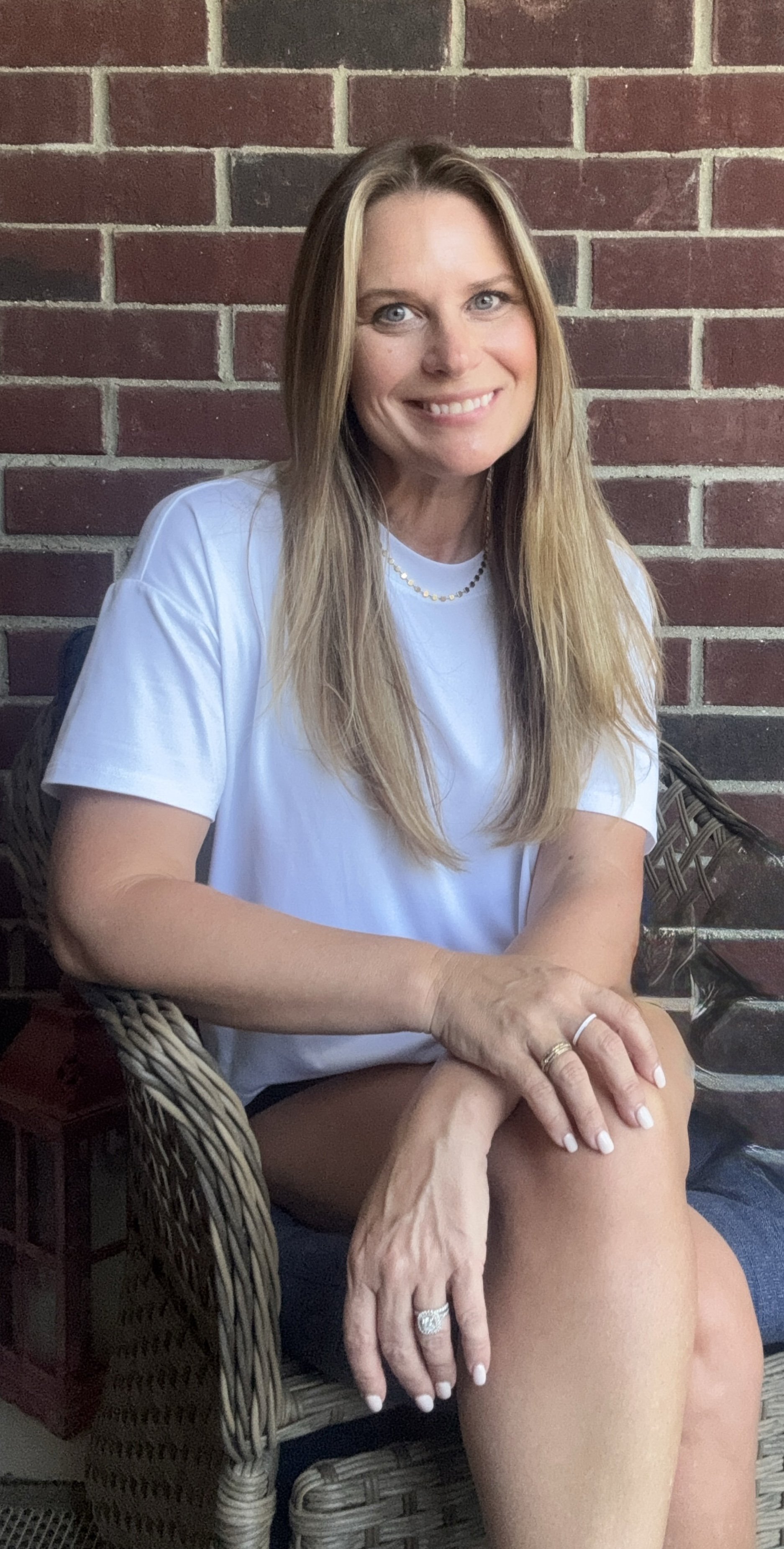 A woman with long blonde hair, wearing a white T-shirt, sitting on a wicker chair in front of a red brick wall, smiling at the camera.