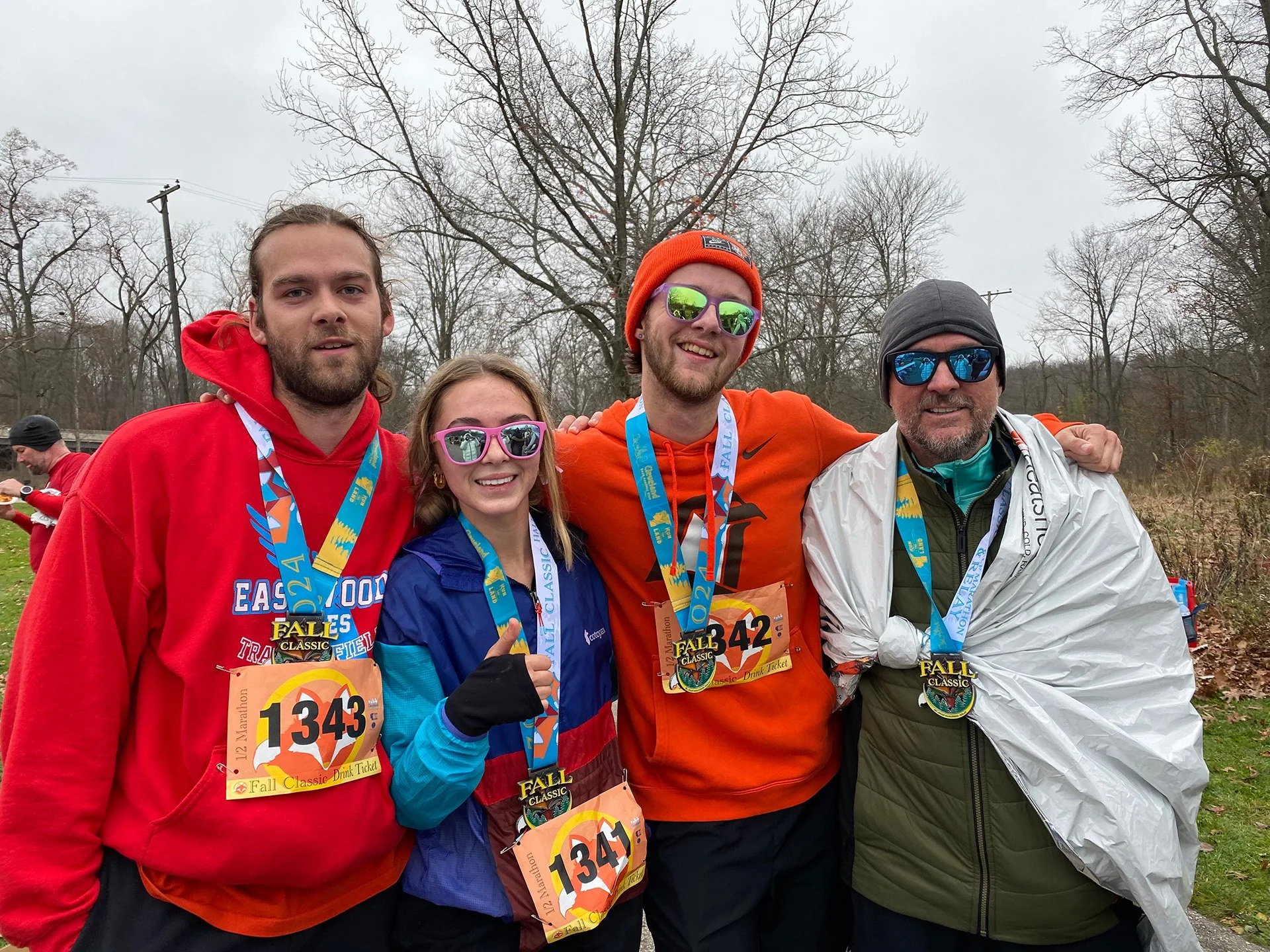 Doug and his children standing outdoors after a race, wearing race medals and bibs. All dressed in athletic gear suitable for cold weather, with trees and overcast sky in the background.