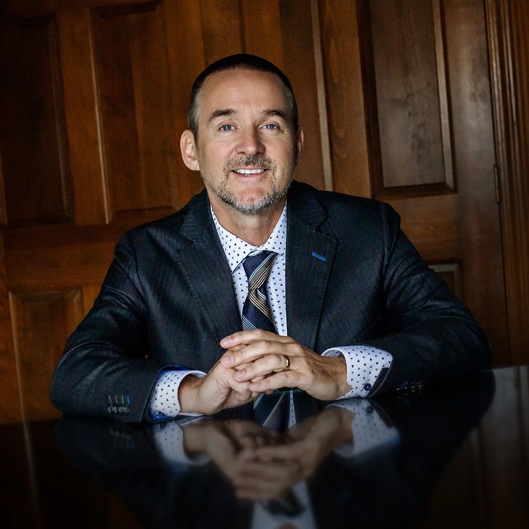 Doug Ruck with short hair and a beard, dressed in a dark suit, white shirt with small dark patterns, and a striped tie, sitting at a reflective black table with hands clasped, in front of a wooden paneled background.