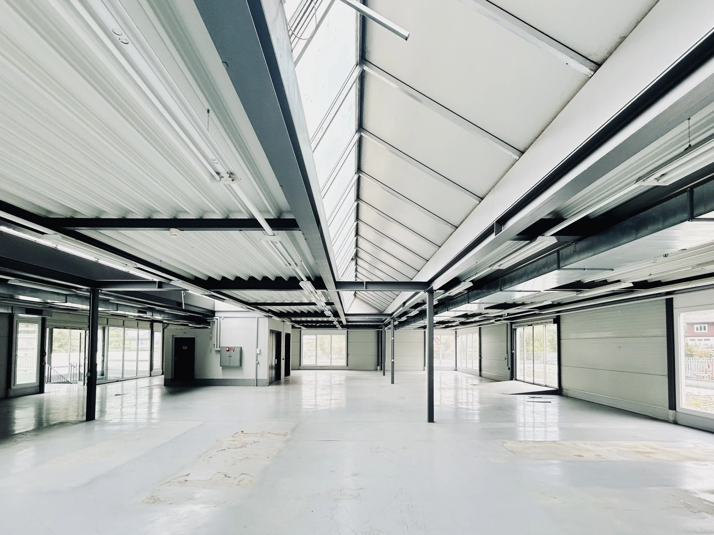 Leerer, heller Raum mit großen Fenstern, Metallstützen und einer geneigten Dachkonstruktion, die für einen Innenraum eines Gebäudes typisch ist.
