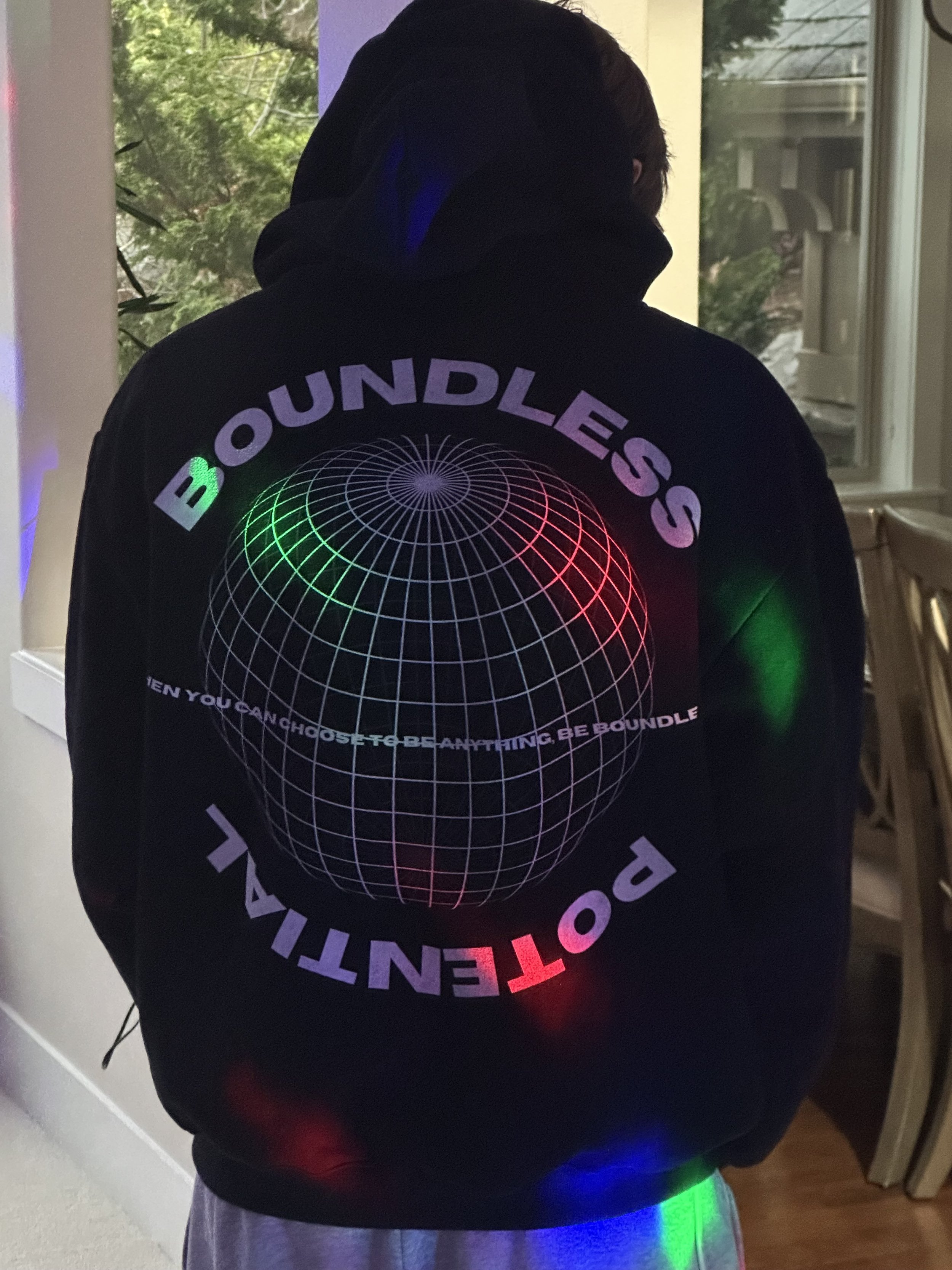 Person wearing a black hoodie with a neon-colored globe graphic and the word 'BOUNDLESS' above and below the globe. The hoodie also has a slogan that says 'THEN YOU CAN CHOOSE TO BE ANYTHING, BE BOUNDLE.' The person is indoors near a window with trees outside.