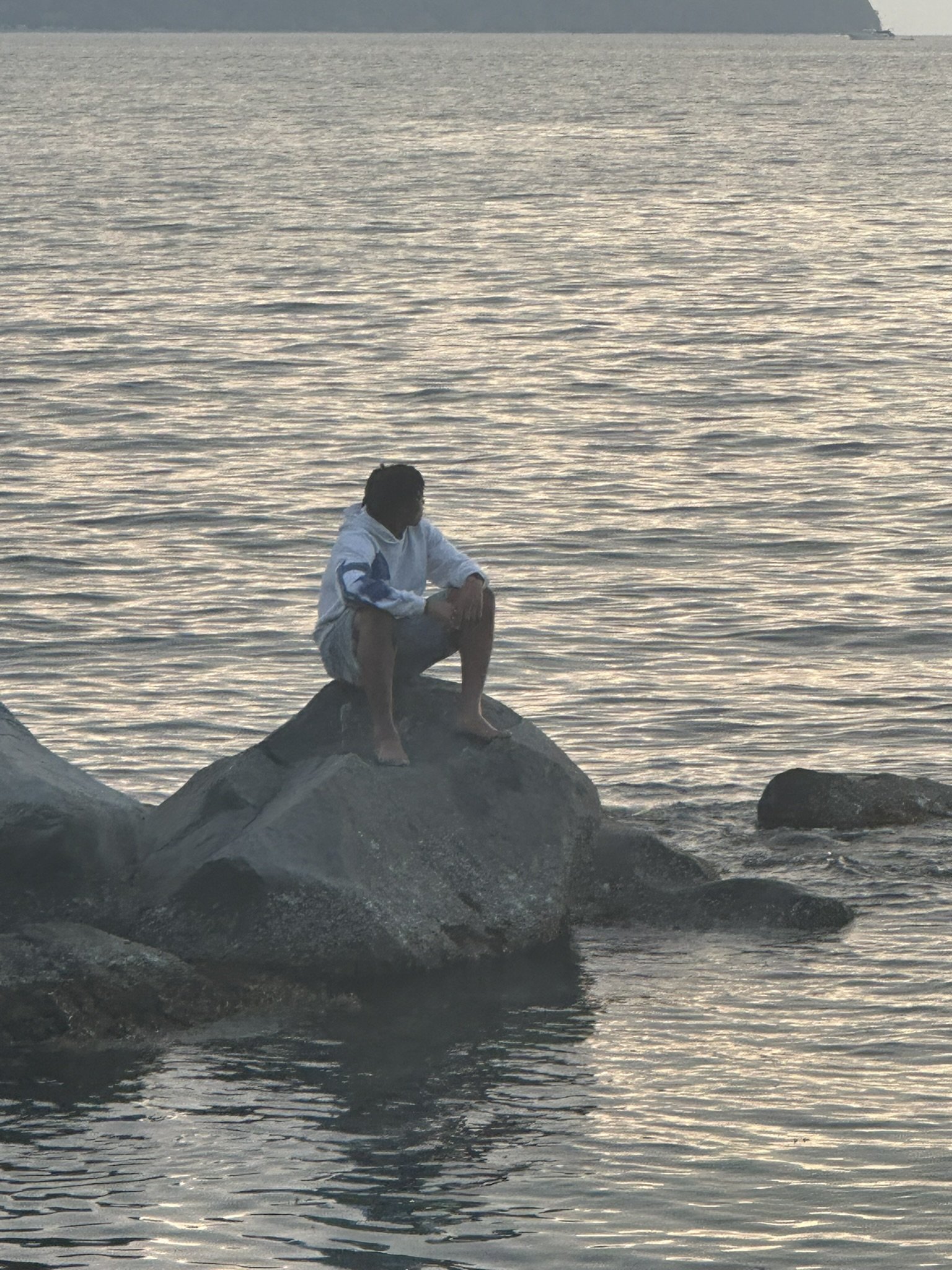 A person sitting on a large rock at the edge of the water, with smaller rocks nearby, during sunset or sunrise.