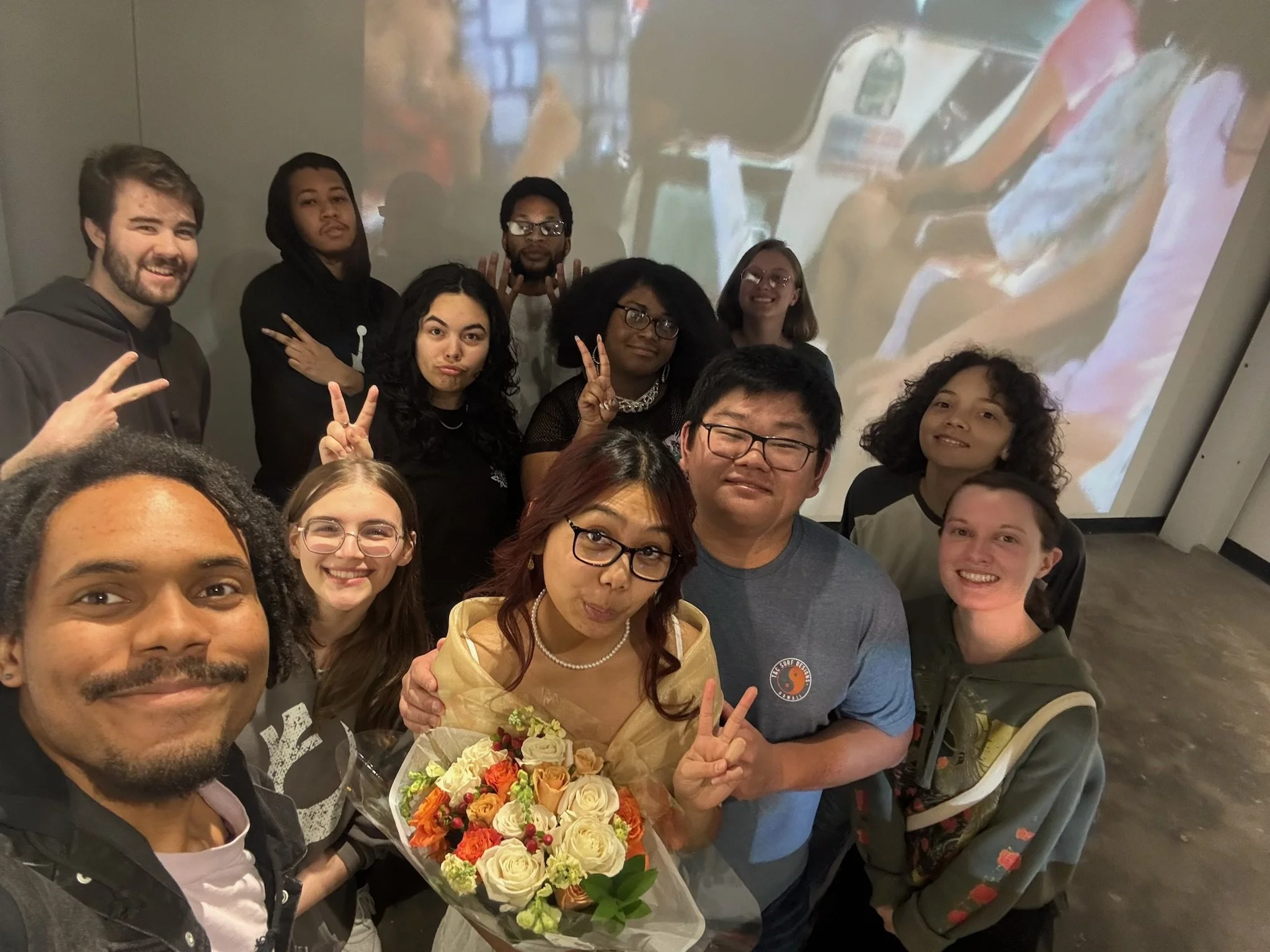 A diverse group of people taking a selfie together, with a woman holding a bouquet of flowers in the center, and a large screen showing an image of seated children in the background.
