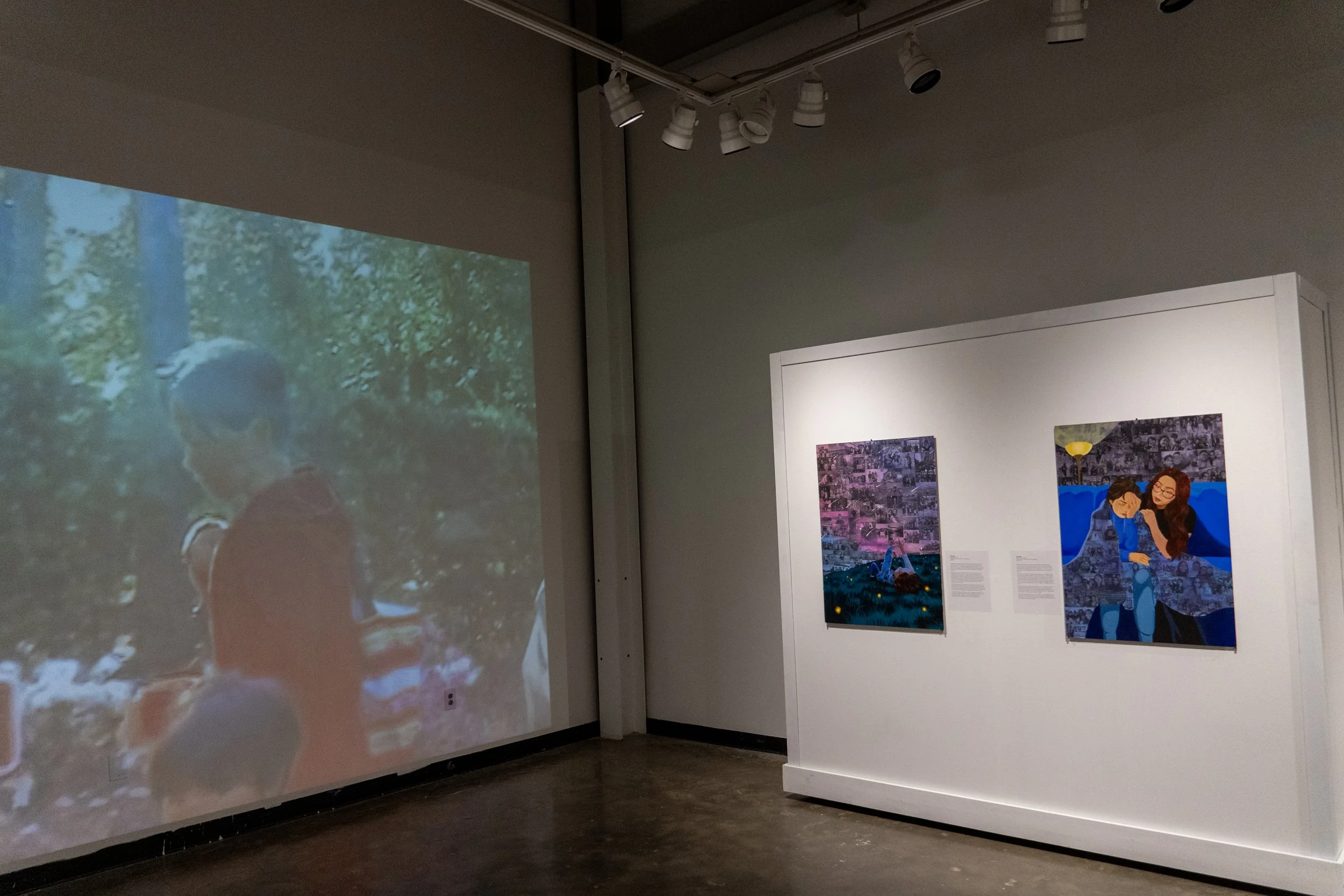An art gallery featuring two paintings on a white wall and a large screen projecting an image of a person in a red outfit outdoors.