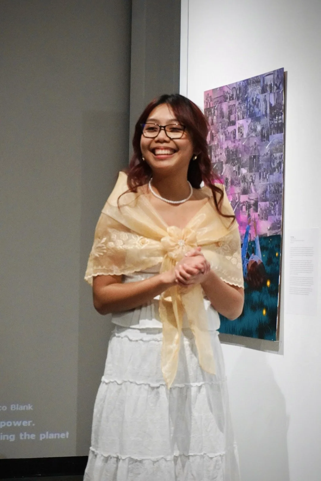 A woman smiling at an art gallery with a collage of photographs on the wall behind her.