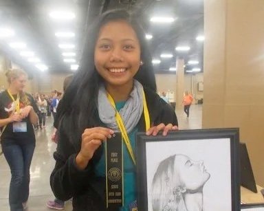 Girl smiling holding a framed pencil drawing and a ribbon in a large indoor event space.