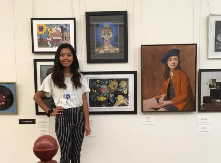 A young woman with long dark hair, wearing a white top and black-and-white striped pants, stands smiling in front of an art gallery wall with framed artwork, including portraits and colorful paintings.