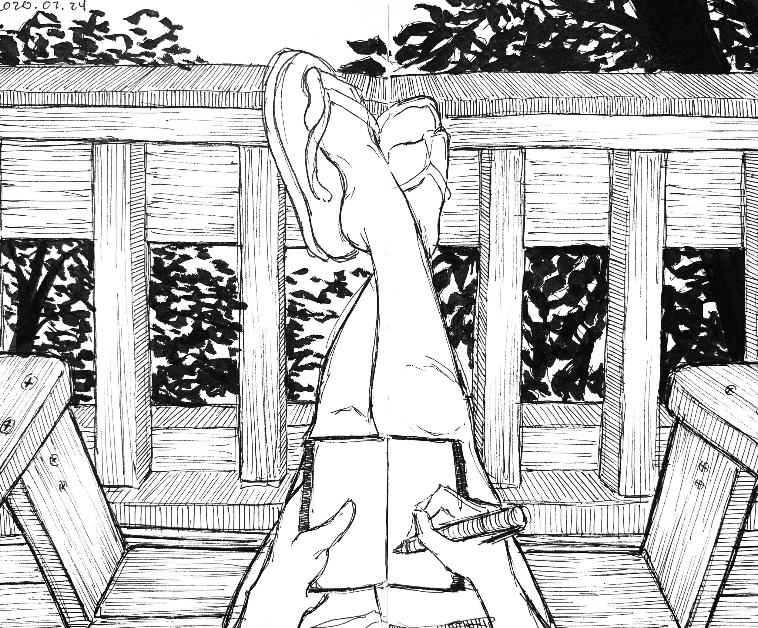 A black-and-white sketch of a person sitting at a wooden outdoor table, holding a pen and writing in a notebook. The scene is viewed from their perspective, with their hands visible in the foreground. There is a wooden railing and leafy trees in the 