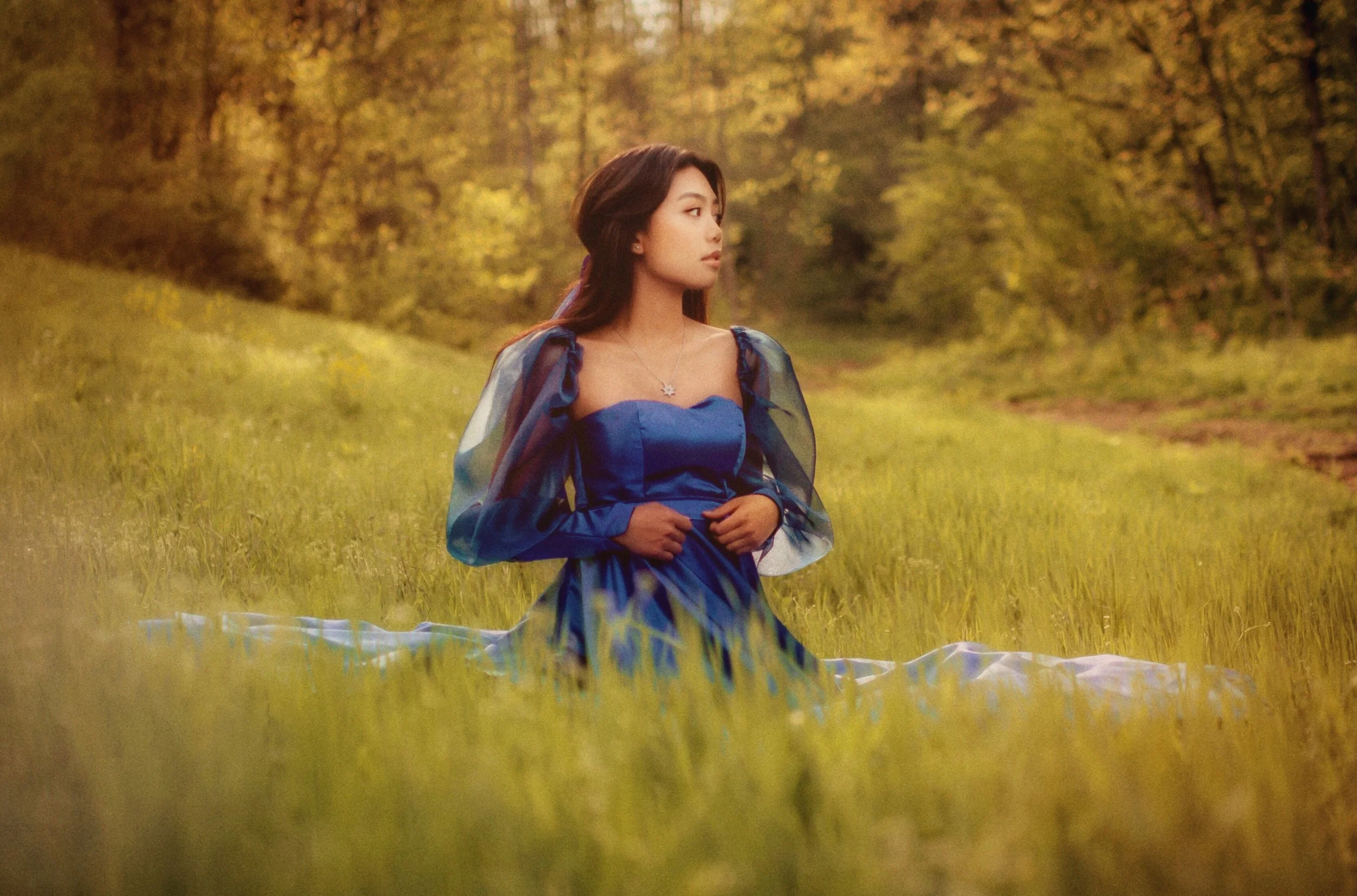 A woman in a blue dress sitting in a grassy field with trees in the background during daylight.