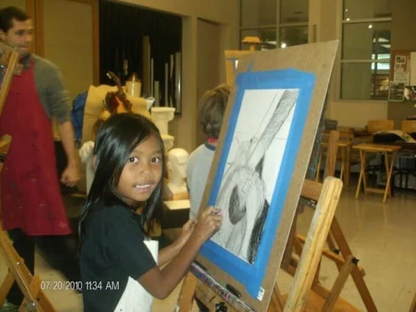A young girl painting on a canvas in an art studio. She is smiling and looking at the camera while working on a black and white artwork."