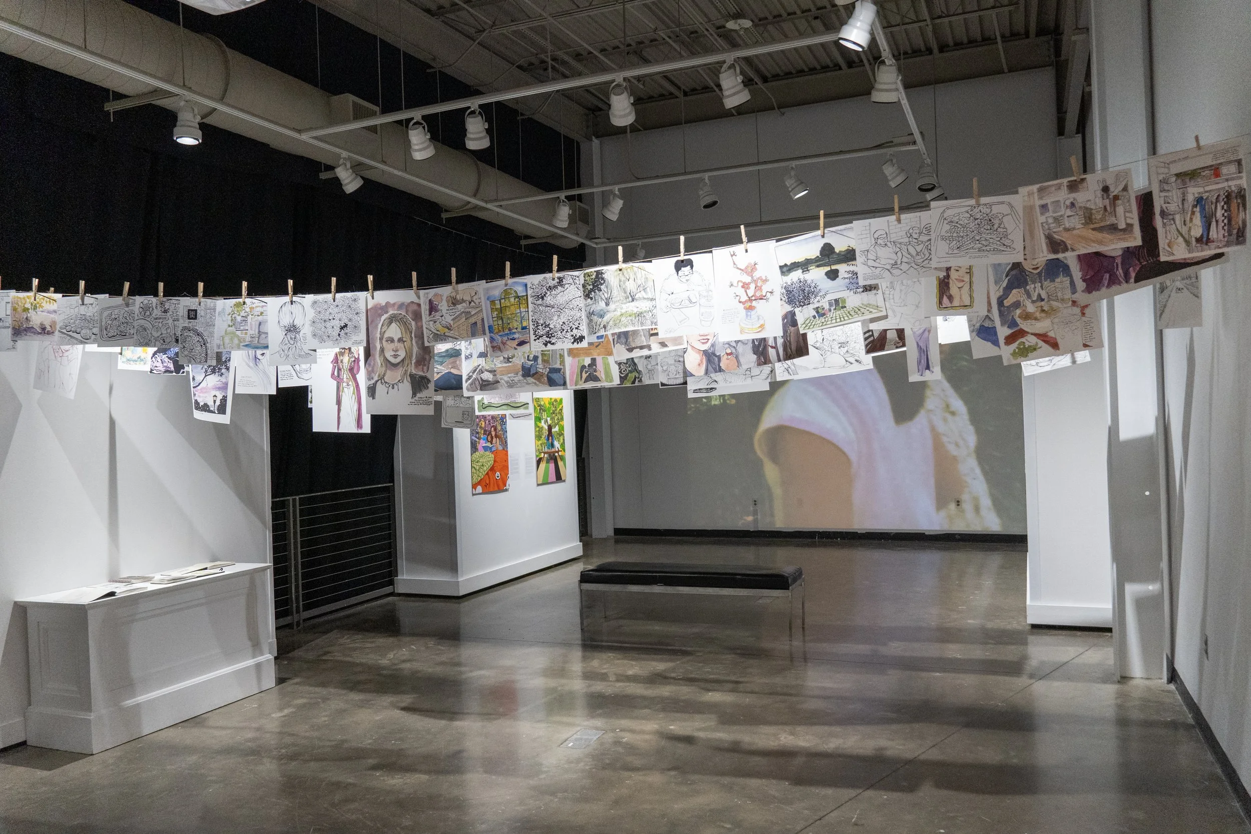 Art exhibition gallery with drawings and paintings hanging on a string with clothespins, and some artworks and a video projection on the wall.