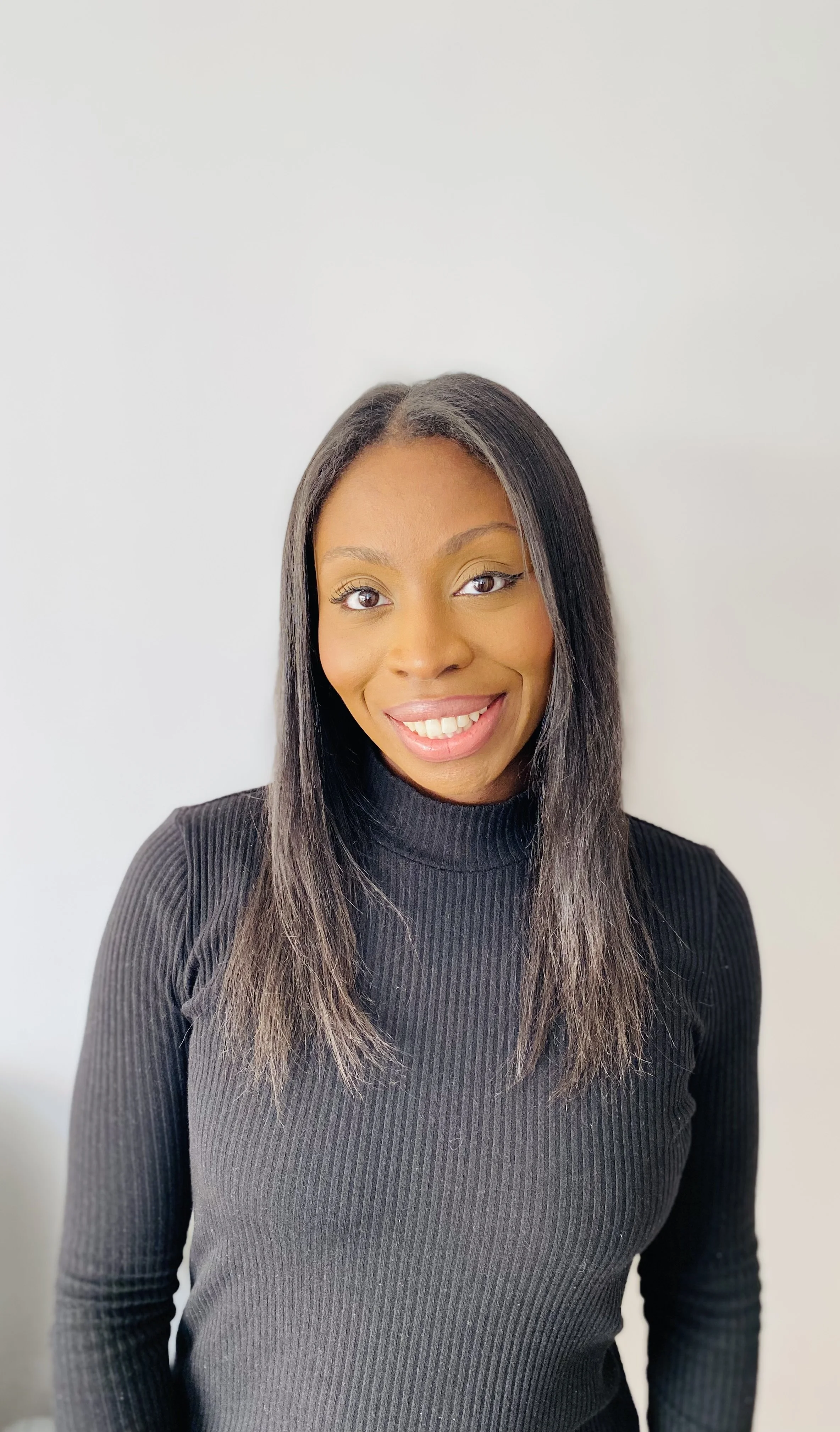 A woman smiling in a black turtleneck with straight, shoulder-length hair, standing against a plain white background.