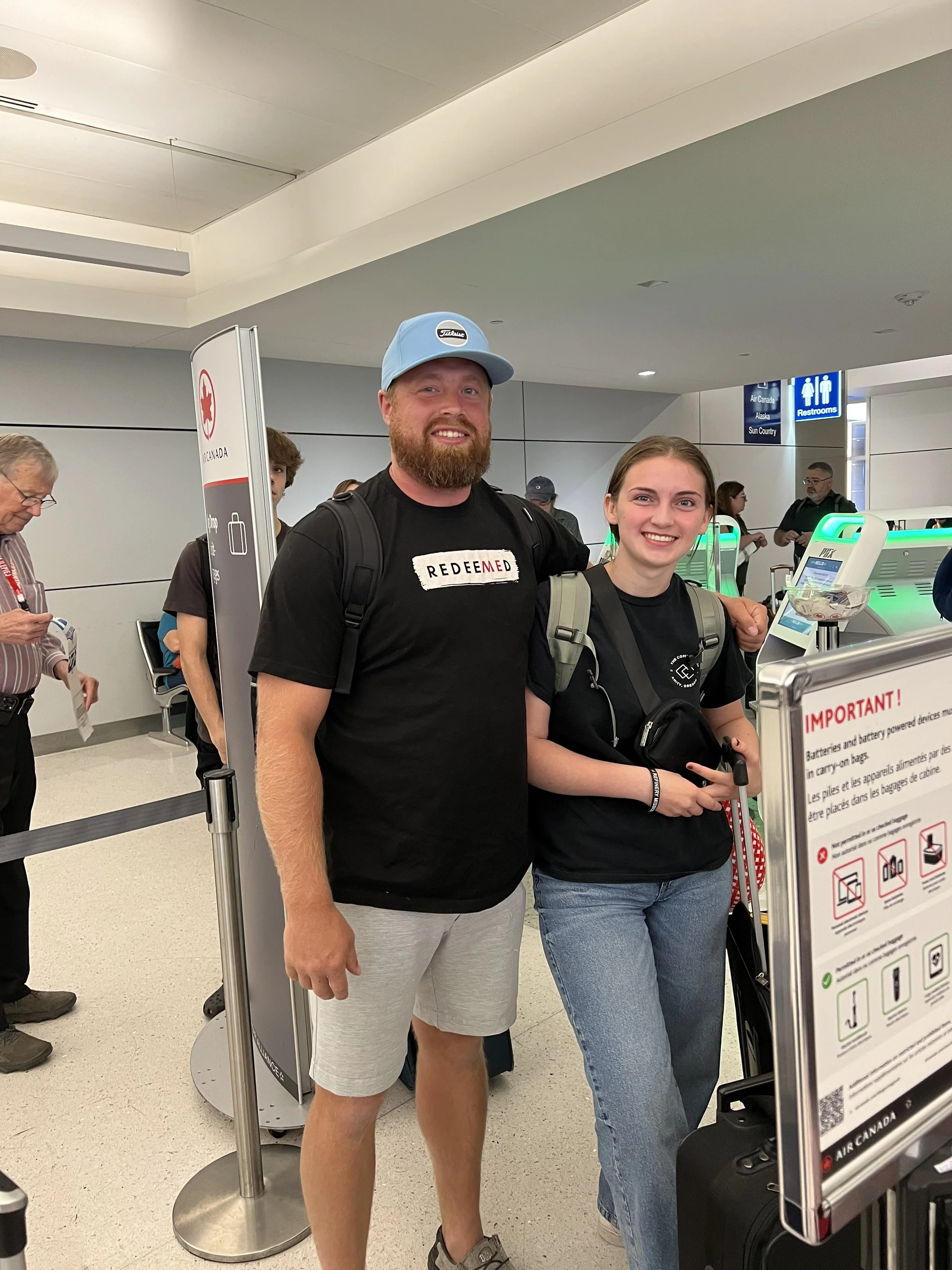 A man and a woman stand in an airport security line, smiling at the camera. The man has a beard, is wearing a light blue cap, a black T-shirt, and light gray shorts. The woman has brown hair tied back, is wearing a black T-shirt, and holds a black ro