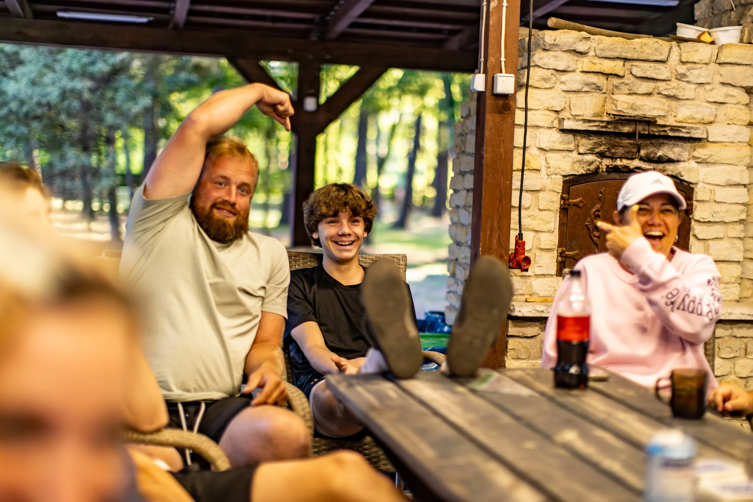 Group of people sitting outdoors, smiling, enjoying a social gathering under a wooden shelter with a stone fireplace in the background.