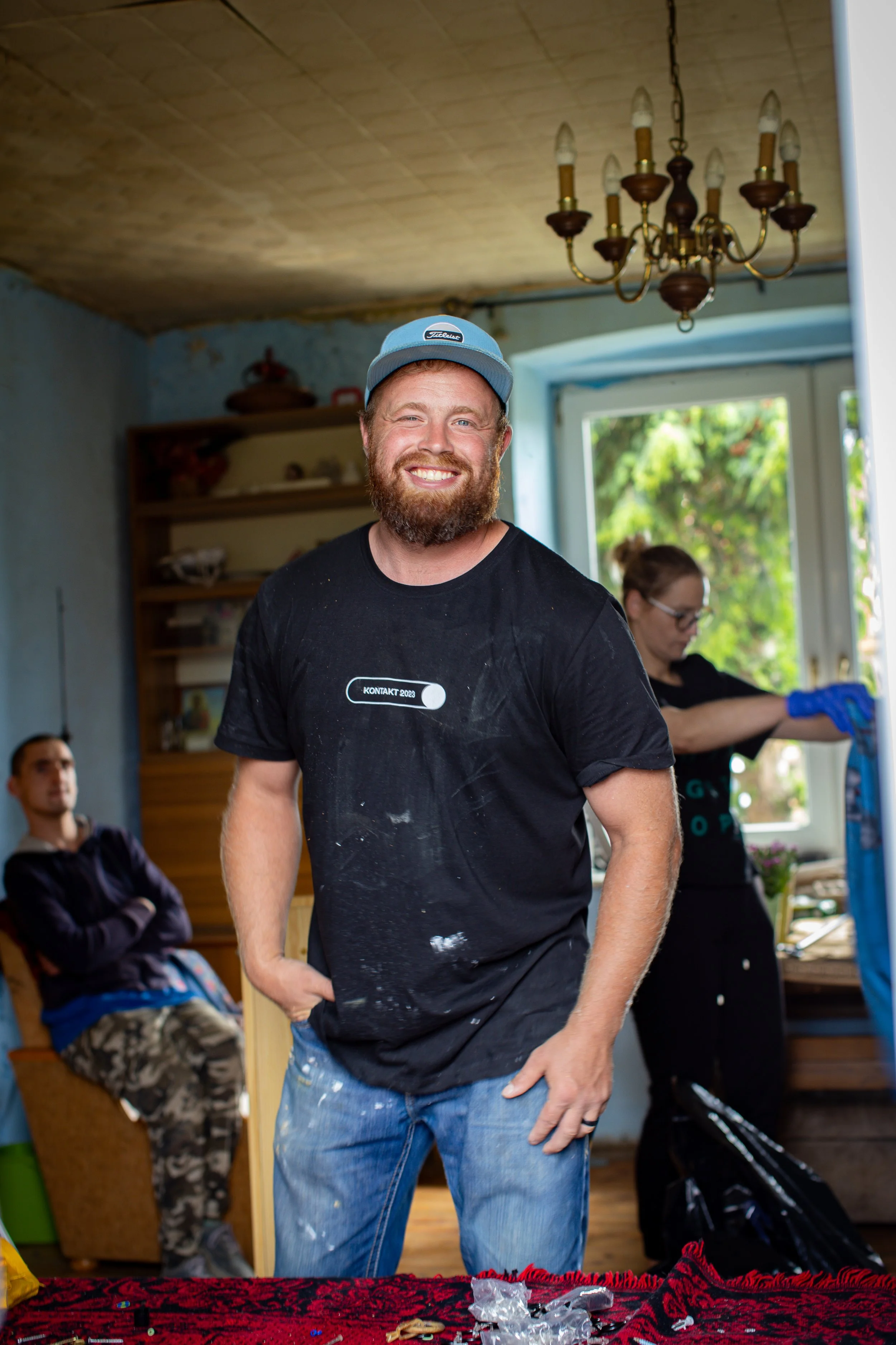 Smiling man with a beard wearing a black t-shirt and a blue cap, standing indoors with people in the background.