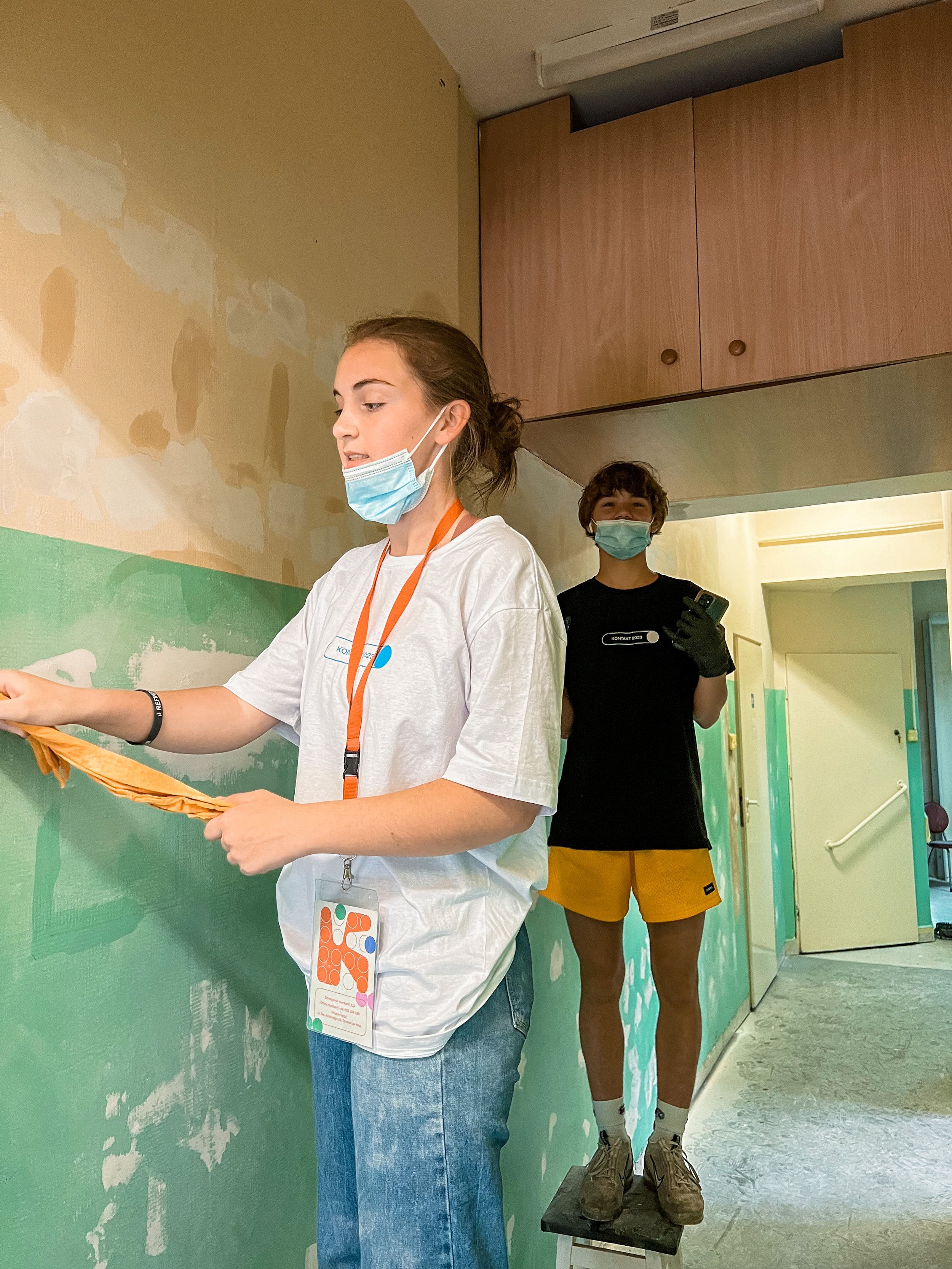 Two people wearing masks working on painting a wall inside a building. One person is on a step stool and the other is holding a paint roller.
