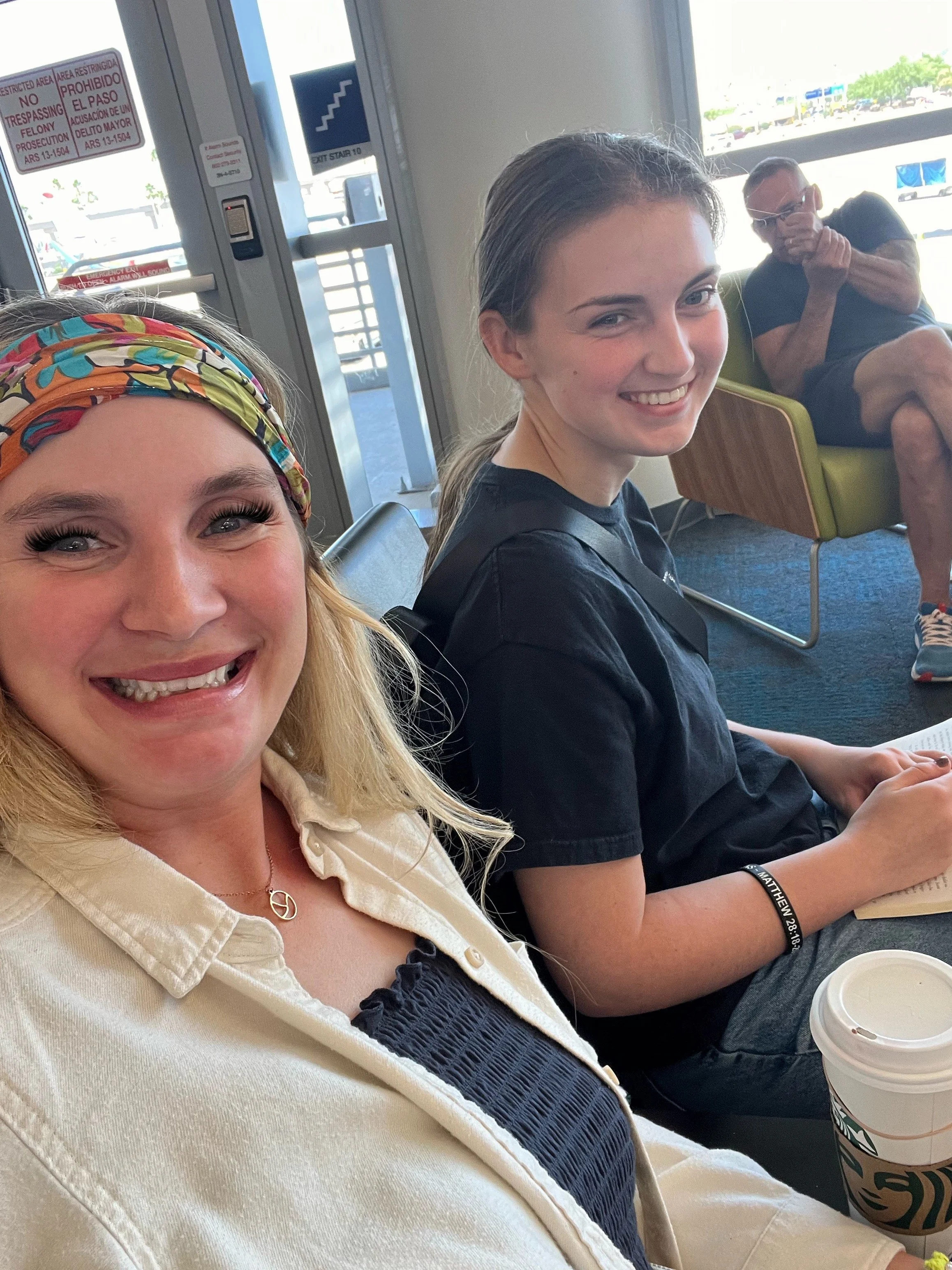Two young women sitting in airport seats, smiling at the camera. One has a colorful headband, the other has a black T-shirt and a bracelet. A man is sitting in the background, looking at his phone.