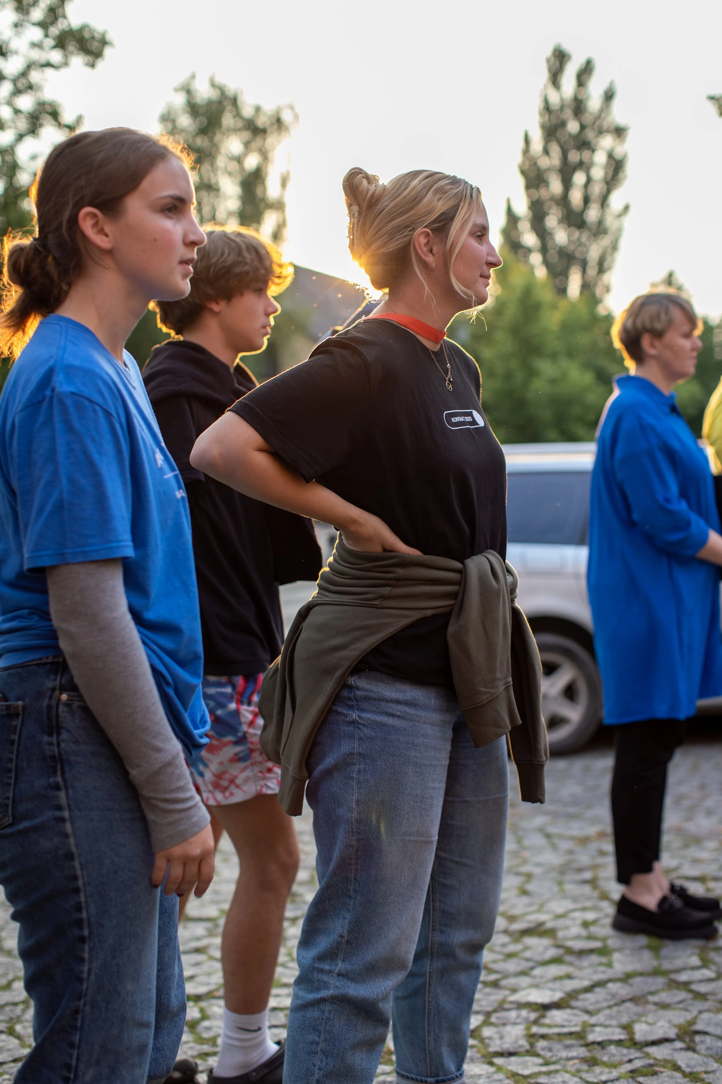 A group of young people standing outdoors on a cobblestone surface during sunset, facing to the right with serious or contemplative expressions.