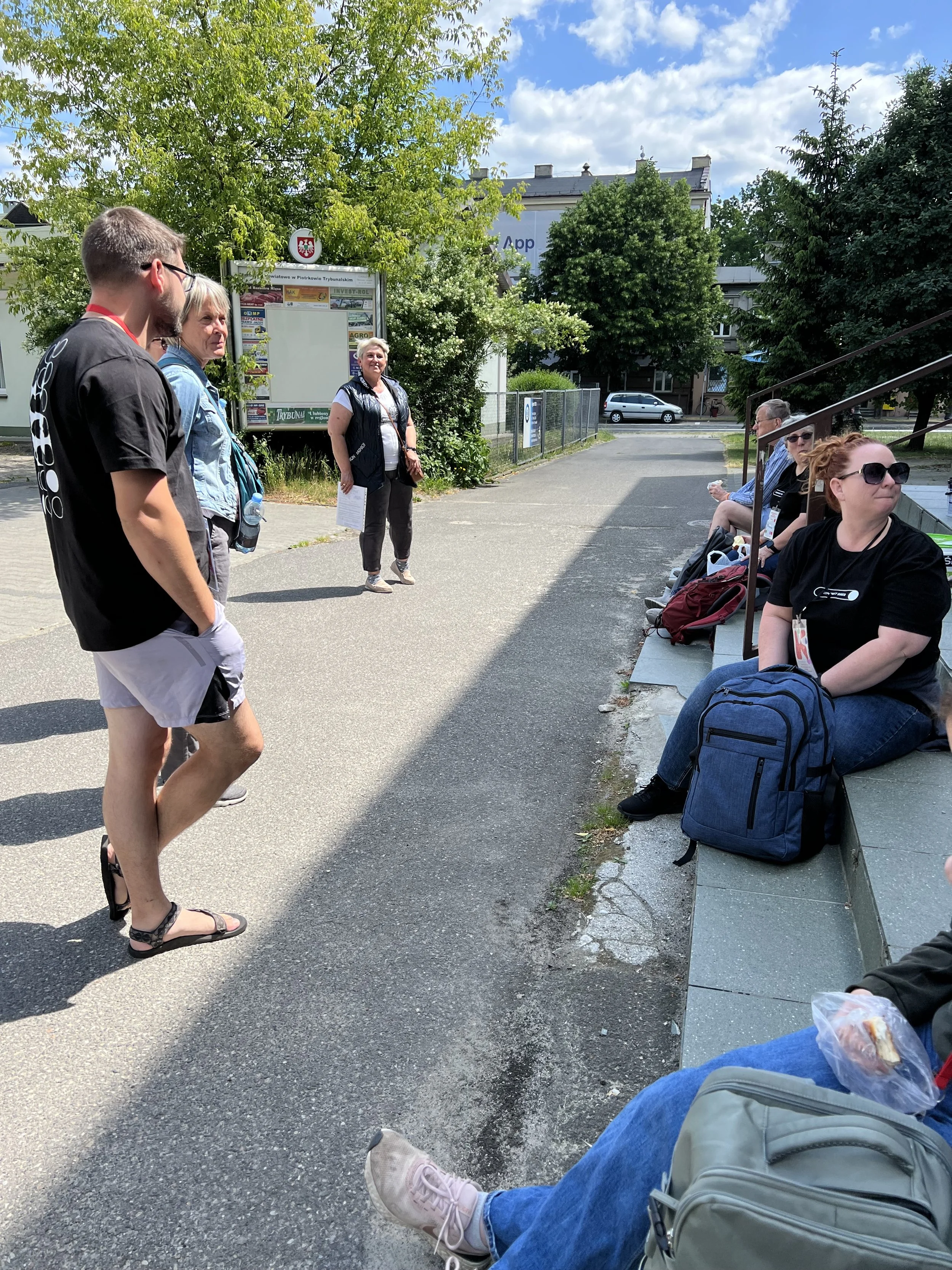 A group of people gathered outdoors in a waiting or meeting area under a partly cloudy sky, with some sitting on benches and others standing, with trees and buildings in the background.