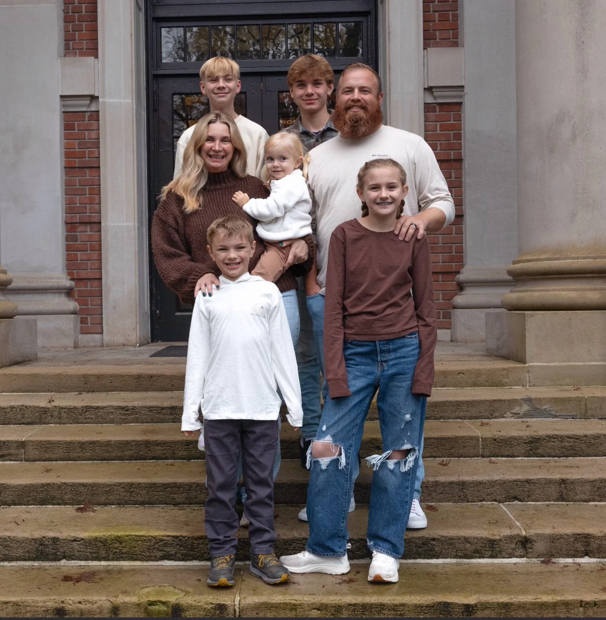 A family of seven standing on the steps in front of a building with a black door, smiling at the camera.