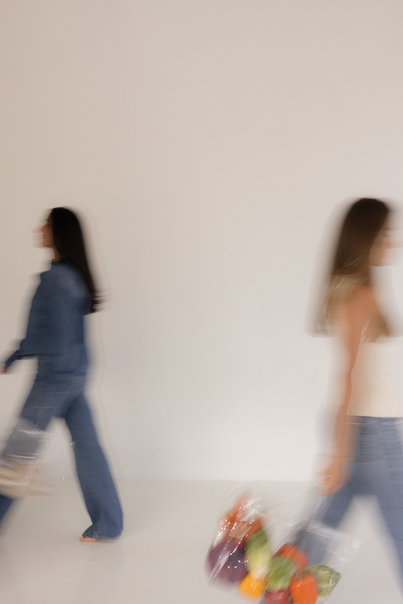 Two women walking in opposite directions with blurred motion, carrying bags, against a plain white wall, with assorted groceries lying on the floor.