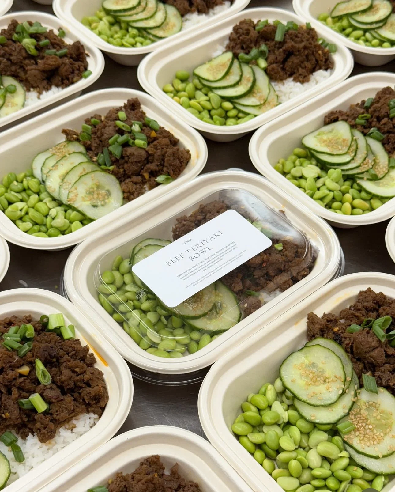 Multiple takeout bowls with beef teriyaki, sliced cucumbers, edamame, and chopped green onions.