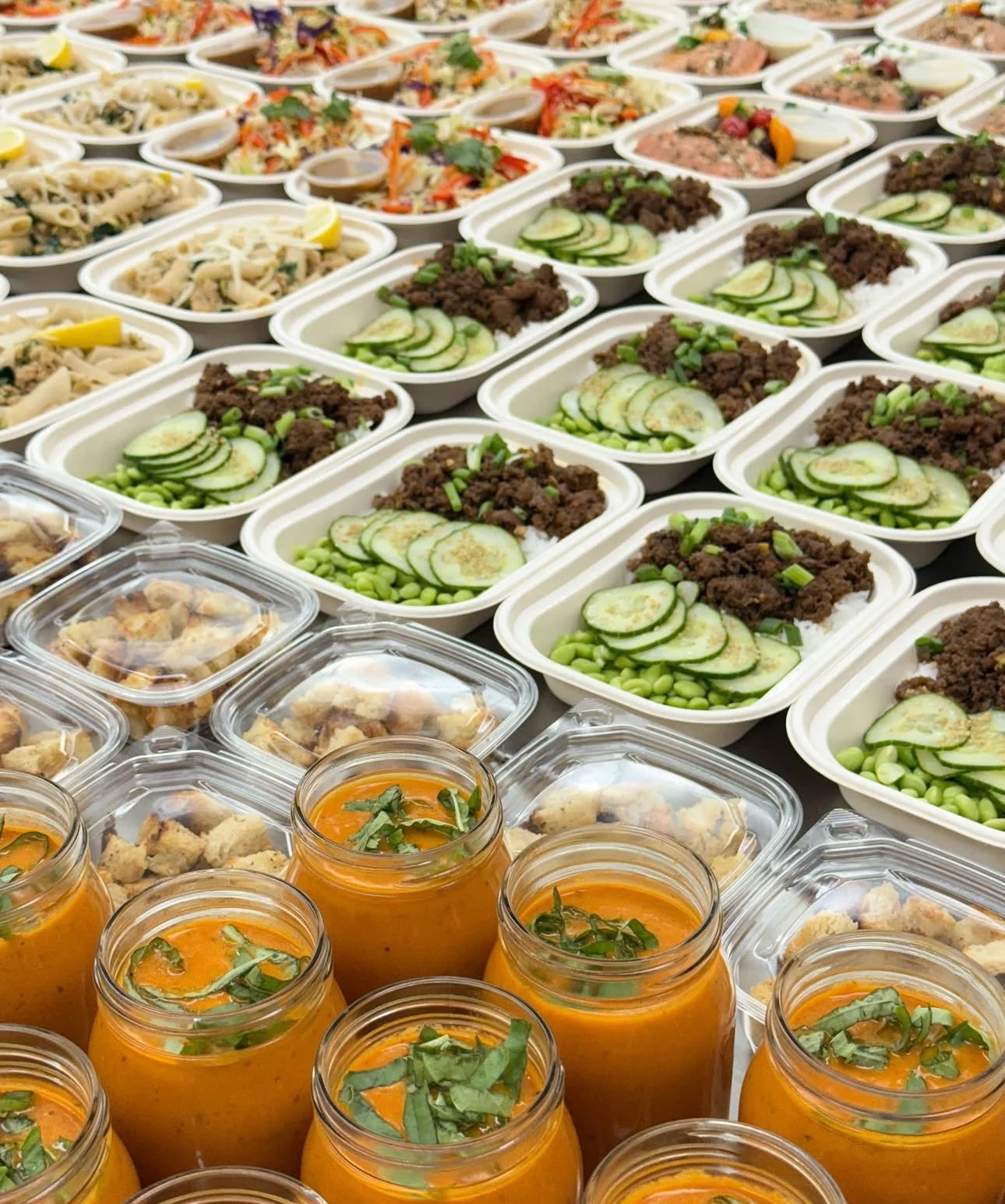Multiple trays of prepared food, including bowls with meat, cucumber slices, and vegetables, as well as jars of orange soup garnished with herbs.