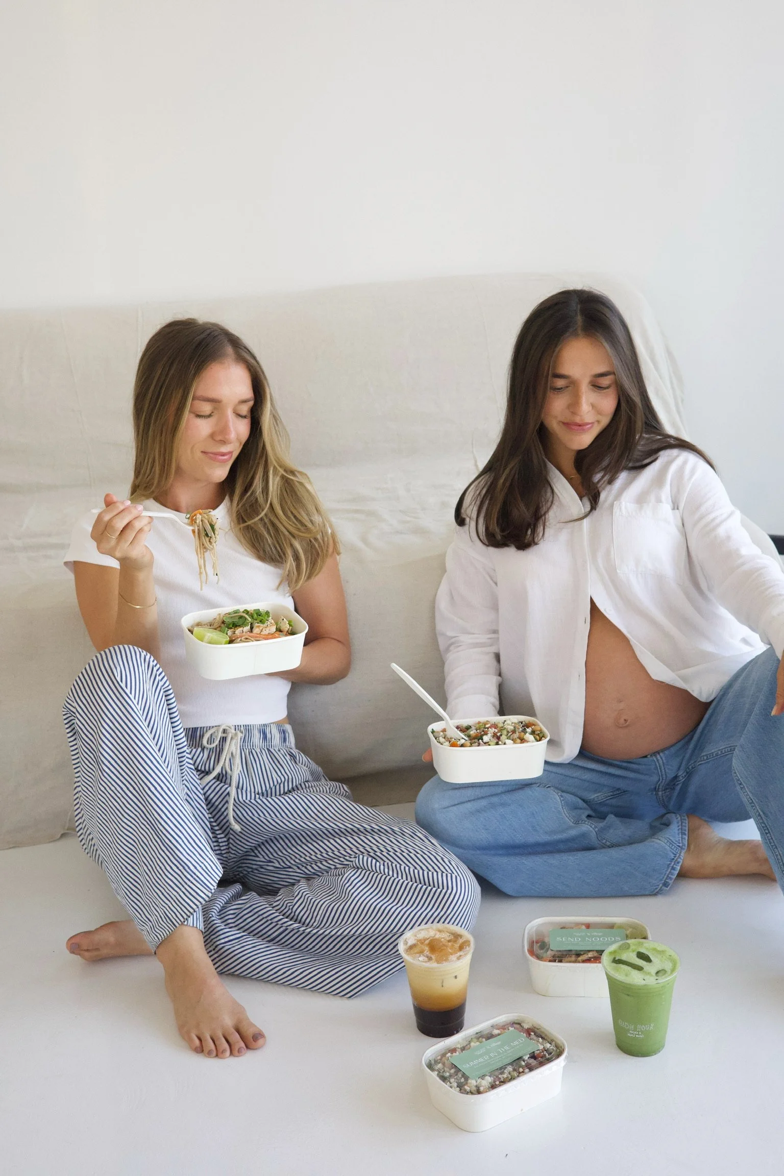 Two women sitting on a white surface, enjoying a meal and drinks together. One woman is holding a bowl of salad, while the other has a bowl with a grain-based dish. There are additional food containers and drinks placed on the surface in front of them.