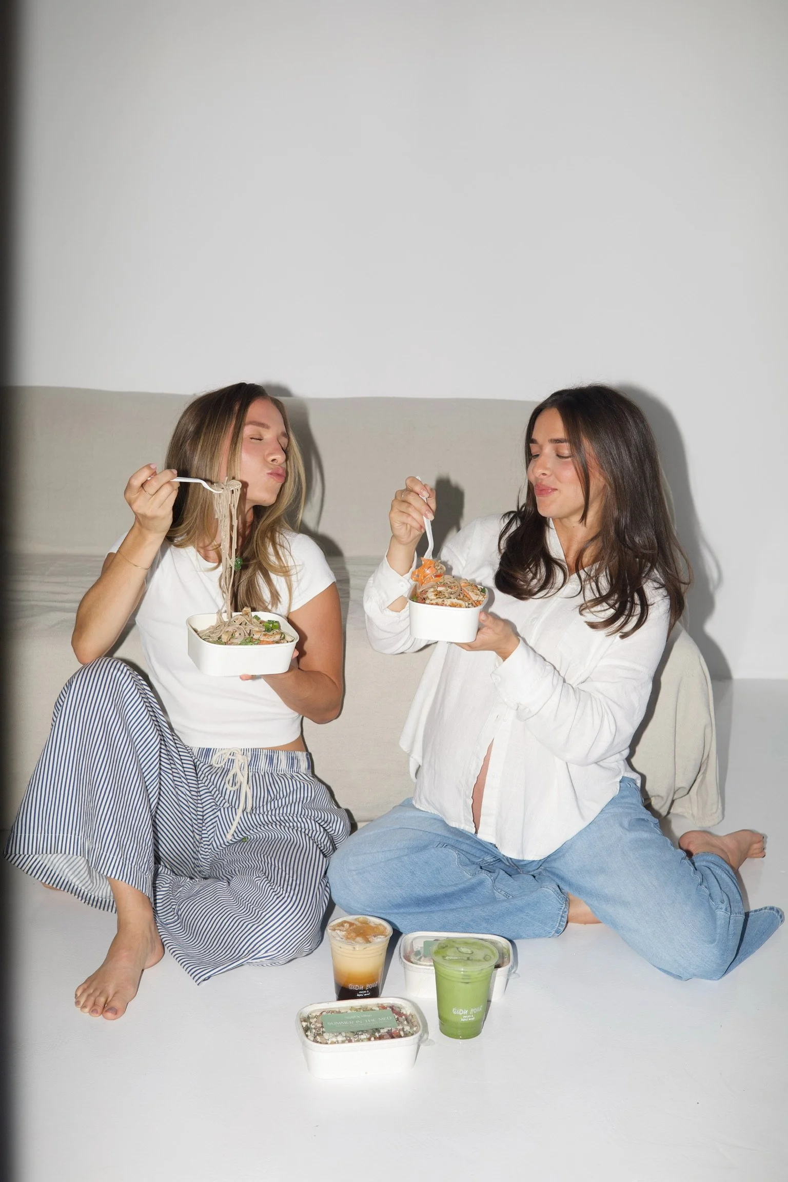 Two women sitting on the floor, eating takeout food with bowls of noodles and other food items around them.