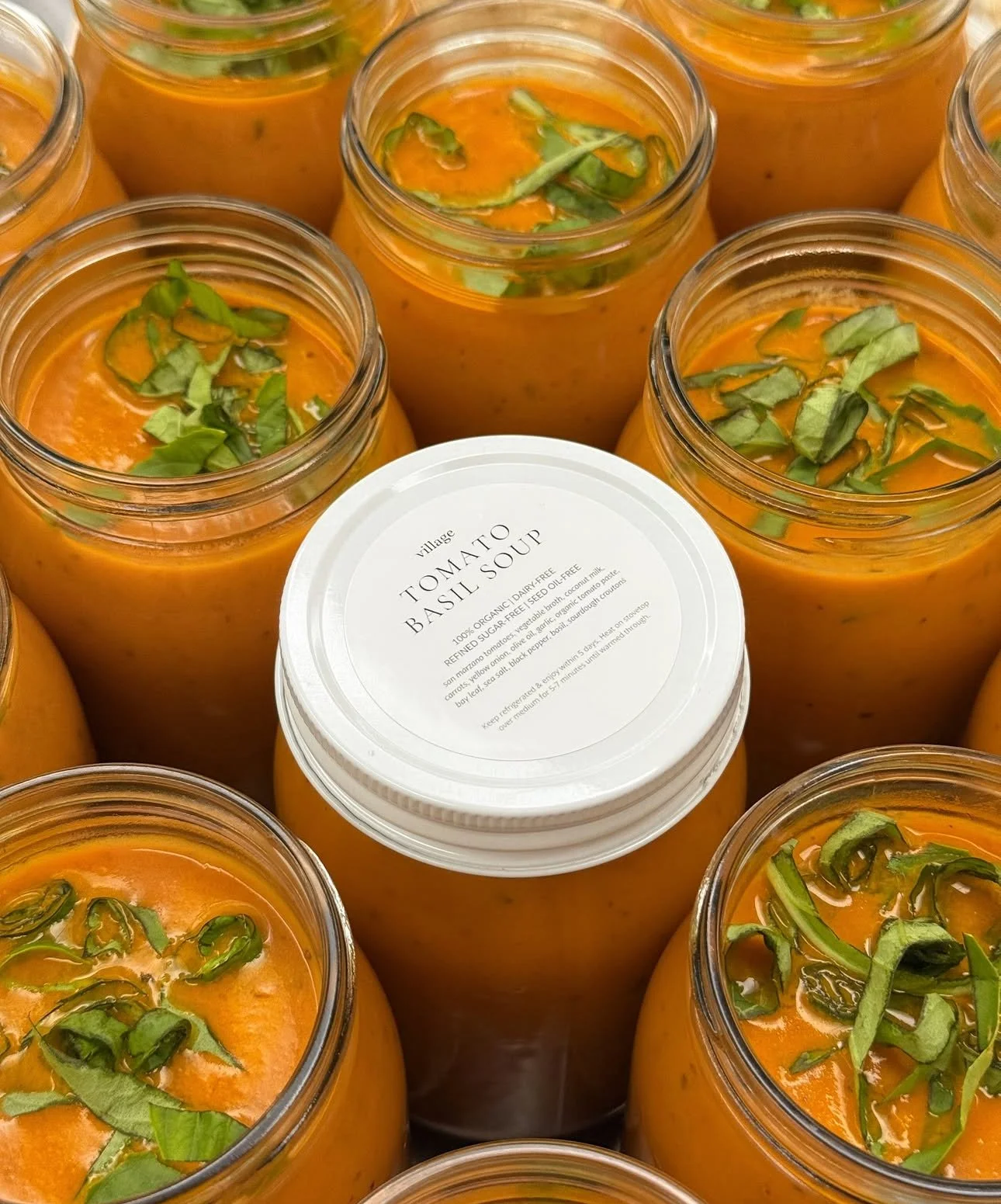 Jars of tomato basil soup garnished with chopped basil leaves, with one jar's lid showing the label 'Village Tomato Basil Soup'.