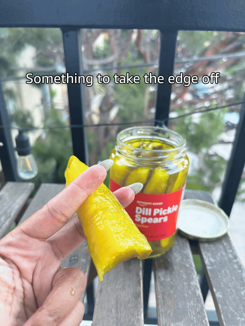 Close-up of a hand holding a bright yellow pickled spear over a wooden surface, with a jar of dill pickle spears and a lid in the background. There is a balcony railing and greenery outside.