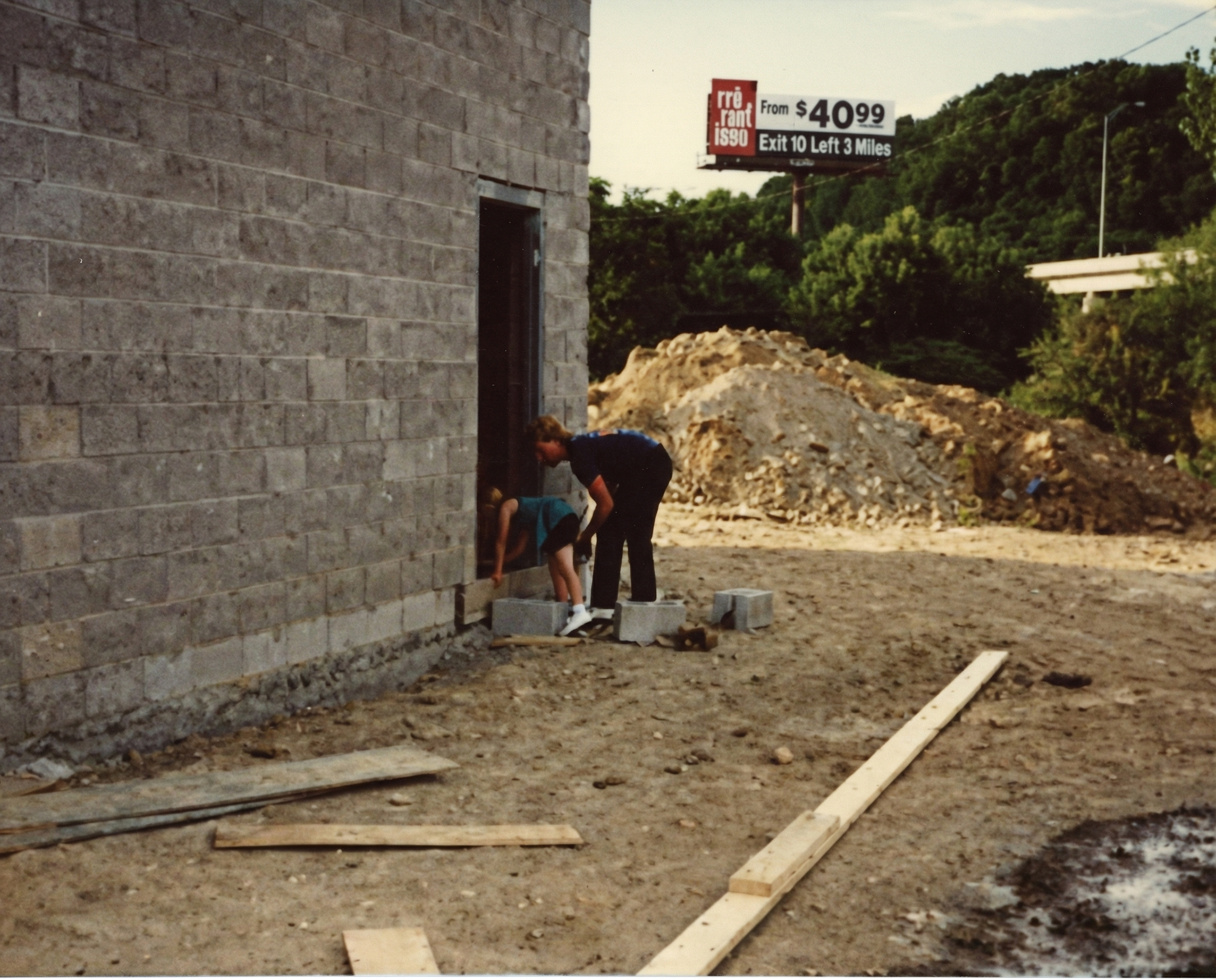 Two people, an adult and a child, working on construction or masonry outside near a building with a brick wall. The adult is helping the child place bricks at the base of the wall. There are piles of dirt and uneven ground, with a wooden plank on the