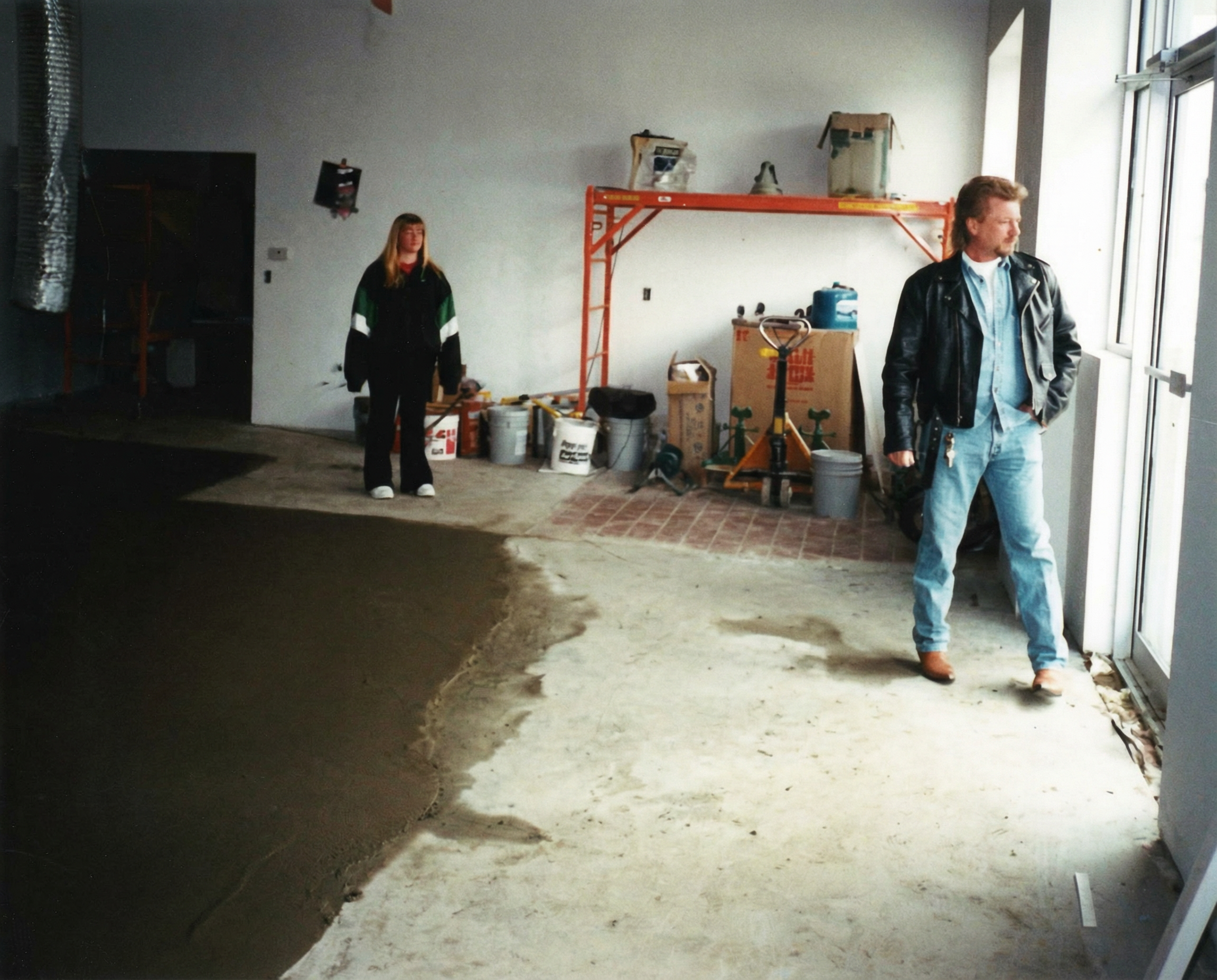 A man and a woman standing inside a room under construction, with construction tools and supplies visible. The man is near the window looking outside, wearing a leather jacket and jeans. The woman stands near the back wall, wearing a black jacket wit