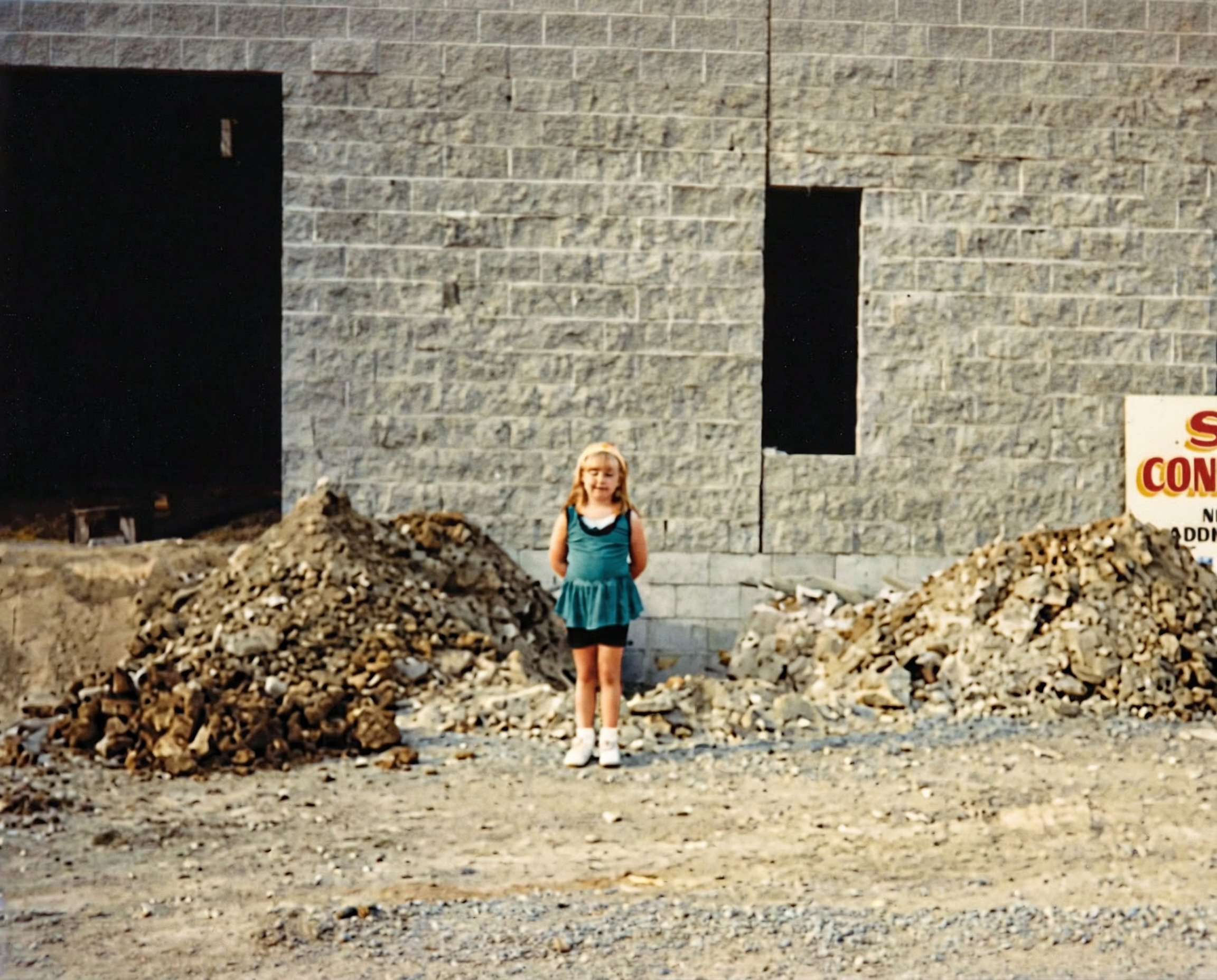 A young girl with blonde hair wearing a teal dress and white sneakers stands in front of a construction site with piles of dirt and a gray brick building under construction.