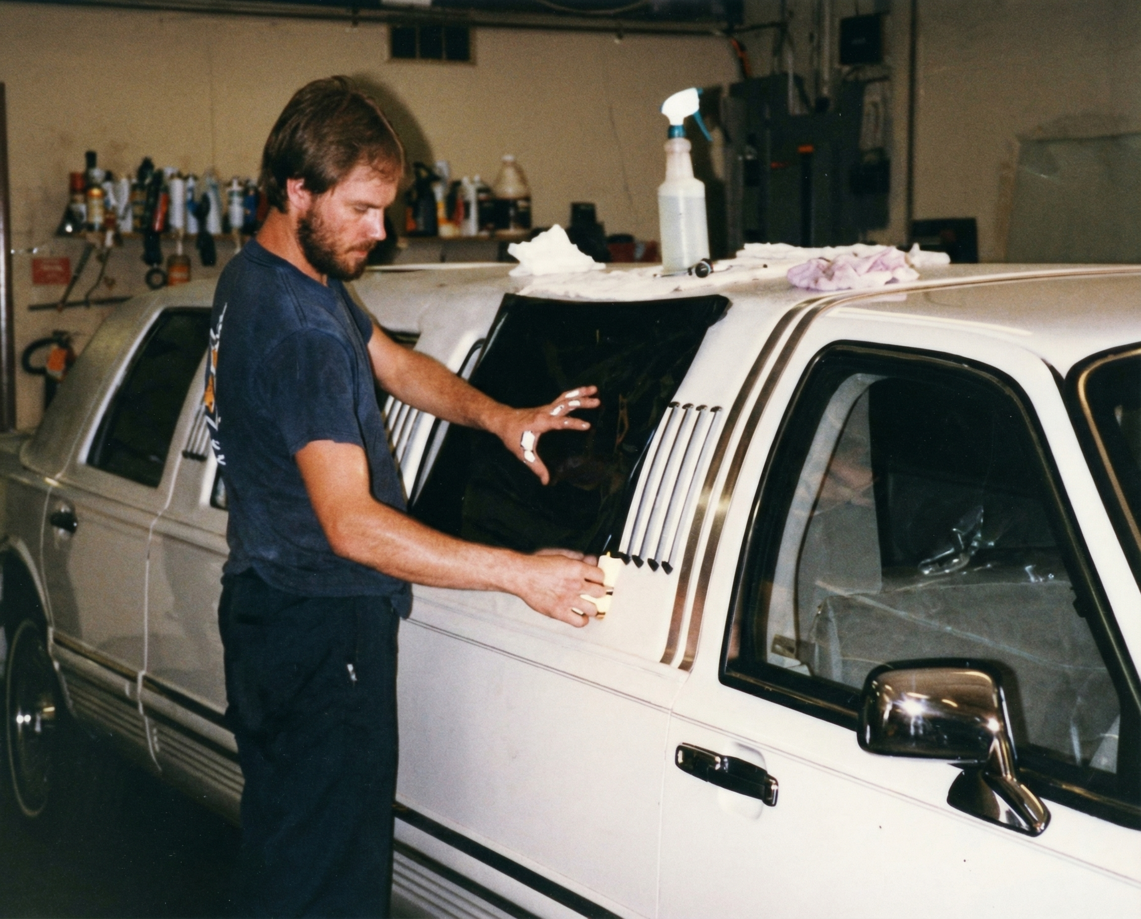 A man working on restoring a white vintage car in a garage. The man is standing beside the car, which has a black windshield. The garage has tools on the wall and supplies on a shelf.