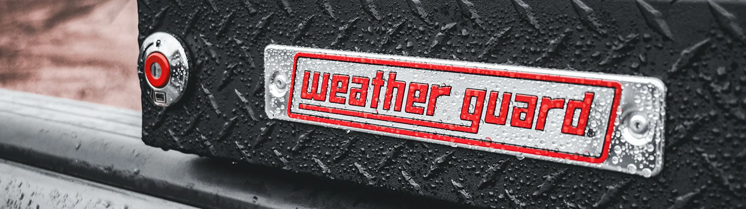 Close-up of a WEATHER GUARD toolbox badge on black diamond-plate truck storage with rain drops, available through Xtreme Car & Truck Accessories in Bridgeville, PA