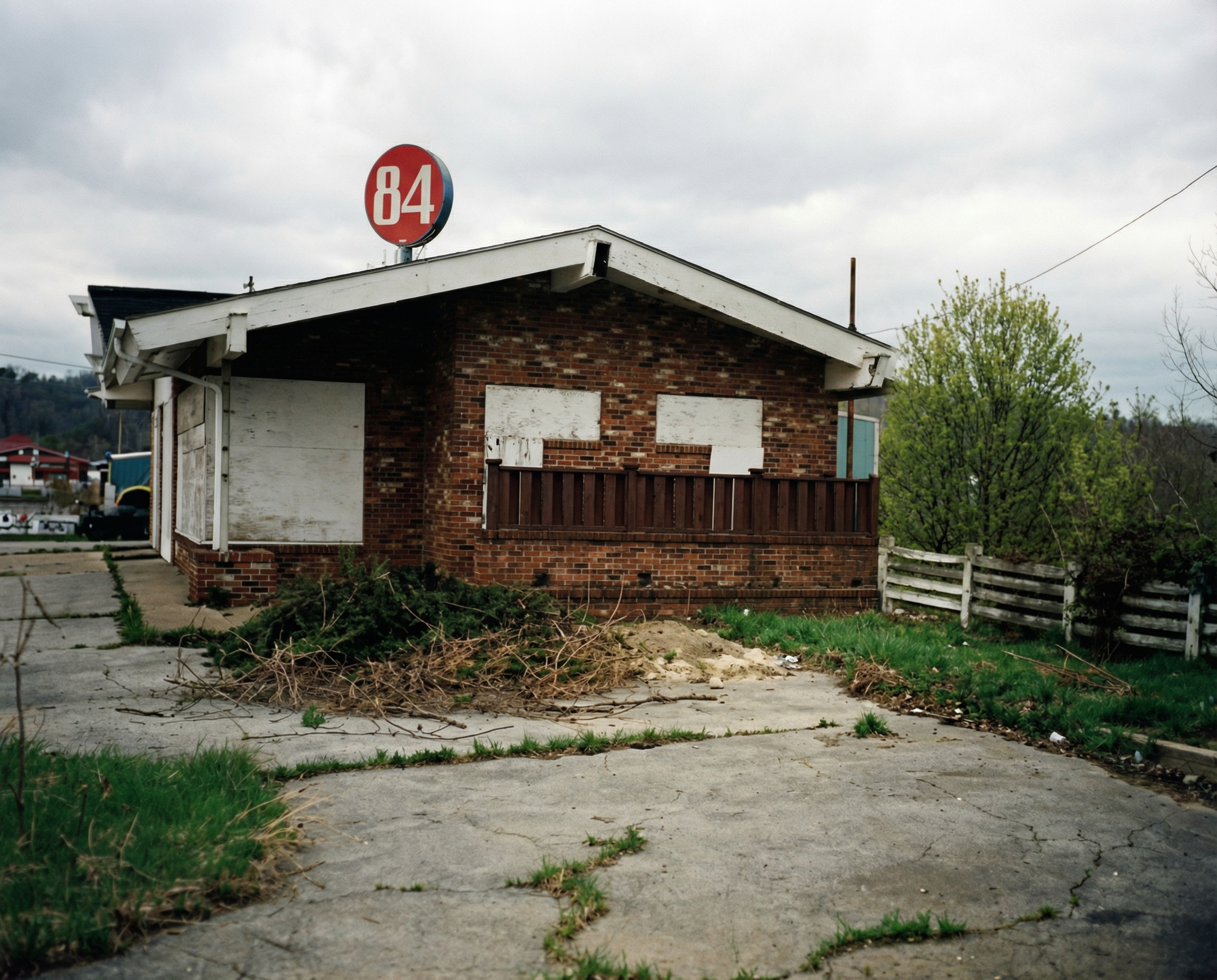 An abandoned, rundown brick building with boarded-up windows and a fenced-in porch. The pavement in front is cracked with weeds growing through, and there are overgrown bushes and trash nearby. A circular sign with the number 84 is on the roof.