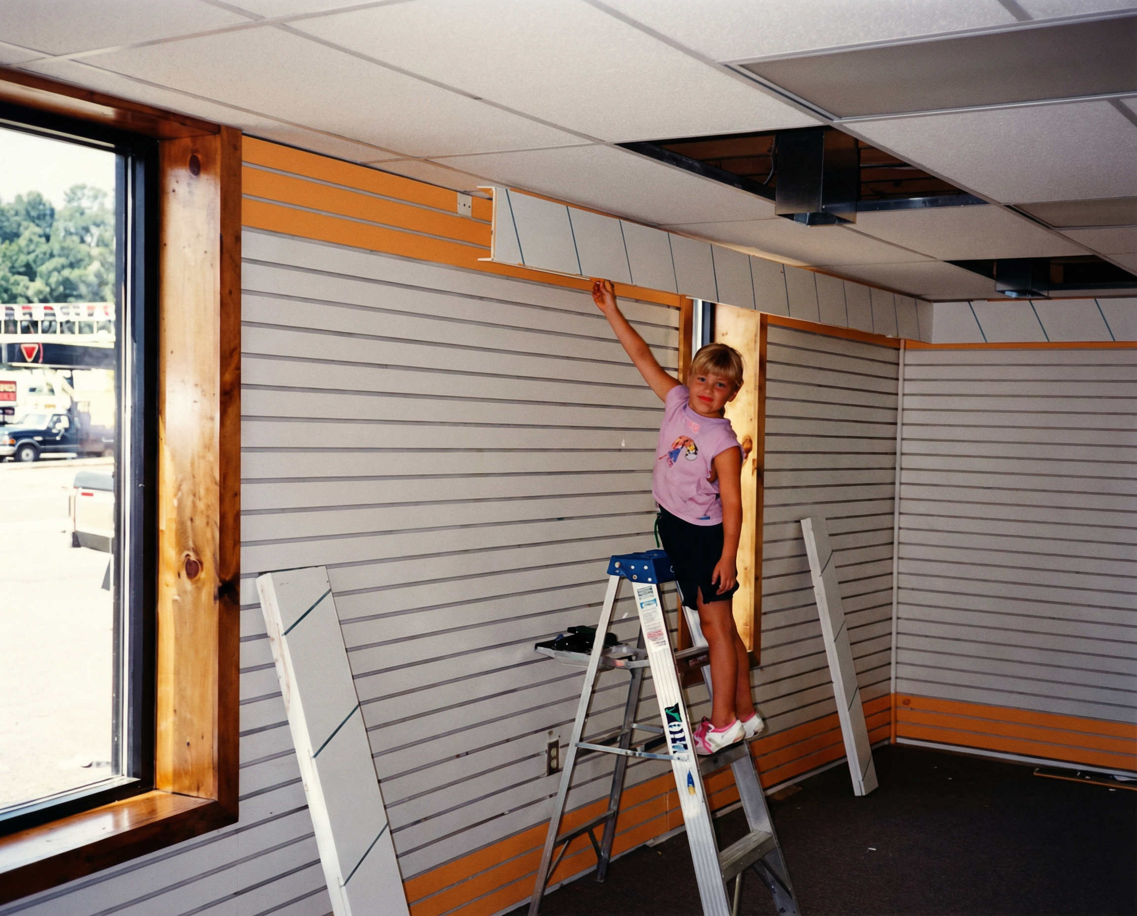 A young girl standing on a step ladder reaching up to install a ceiling tile in a store or office renovation. The room has slatwall panels being installed, with some drywall or insulation visible in the ceiling.