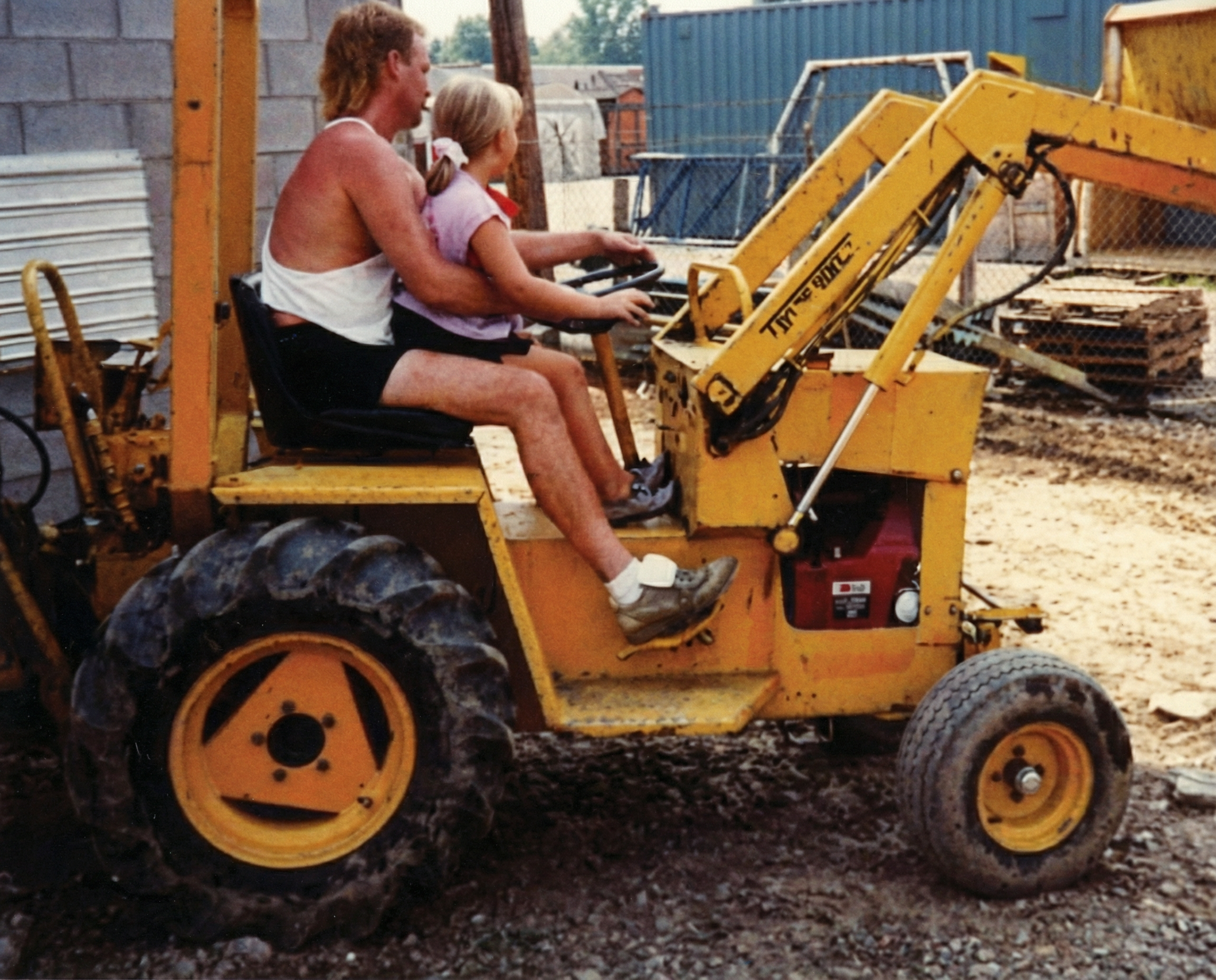 A man and a young girl sit on a small yellow construction vehicle, with the man guiding the girl as she operates the vehicle at a construction site.