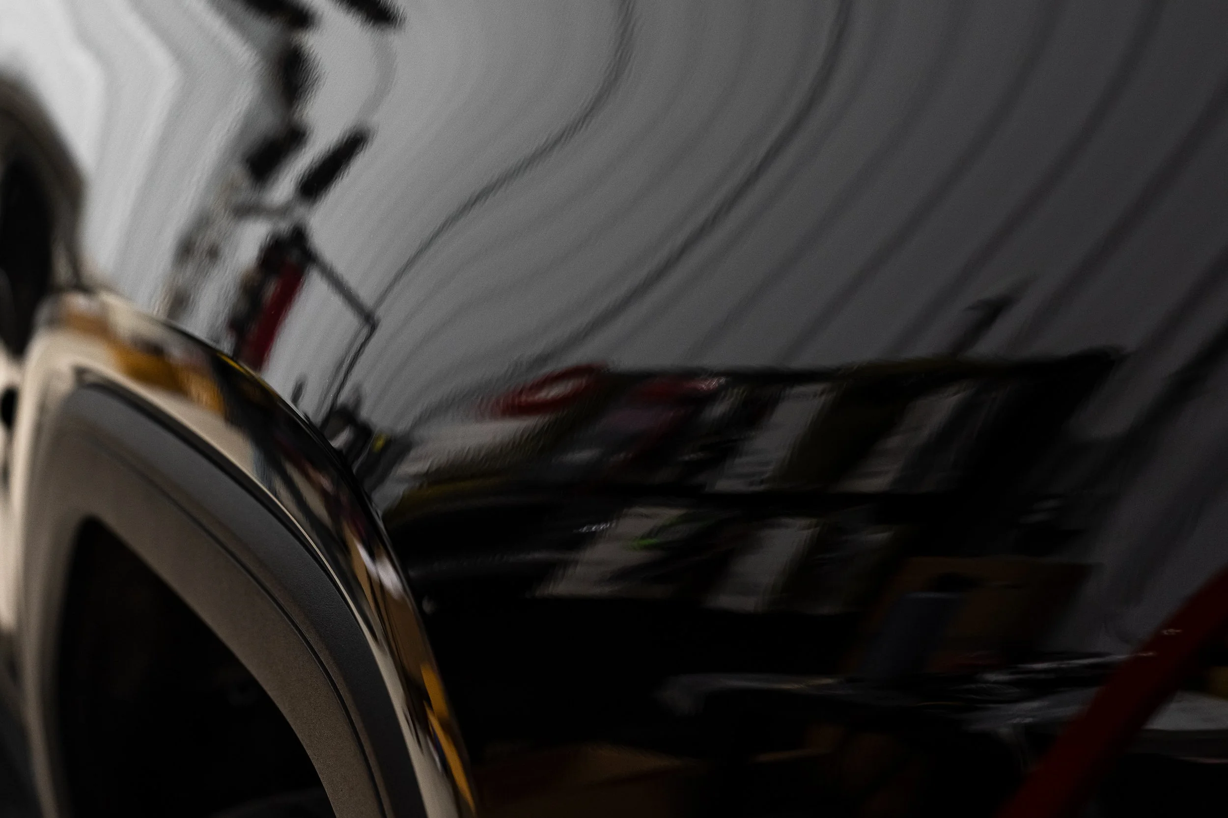 A close-up of a black shiny car with its reflection visible in the paint, showing part of the wheel and body of the vehicle.