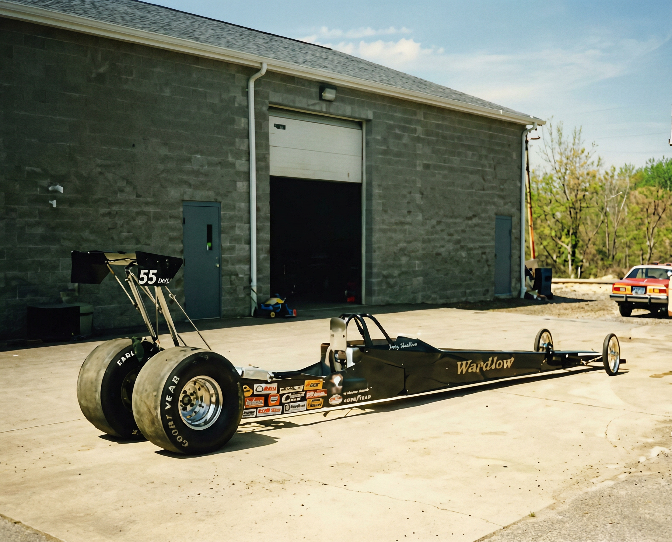 A black drag racing car with the name 'Wardlow' on the side, positioned outside a gray garage building on a concrete driveway. The car has large rear tires and a sleek, elongated body. There are racing sponsor stickers on the front of the car.