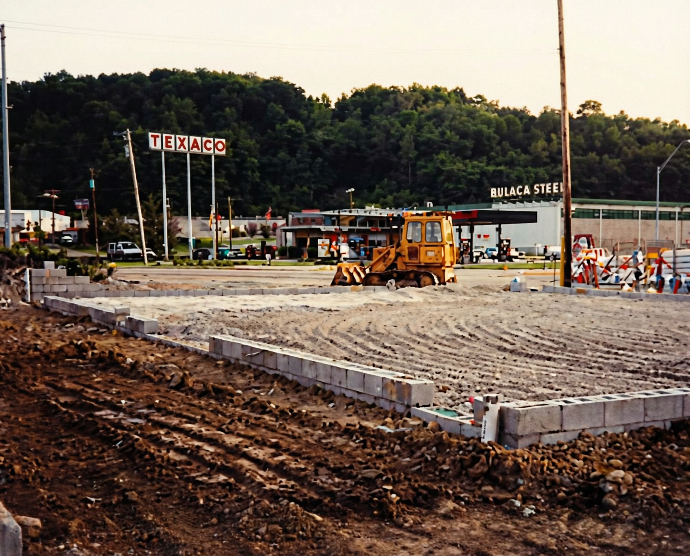 Construction site with a bulldozer working on a sidewalk, with traffic and Texas sign in the background, and a hill covered with trees.