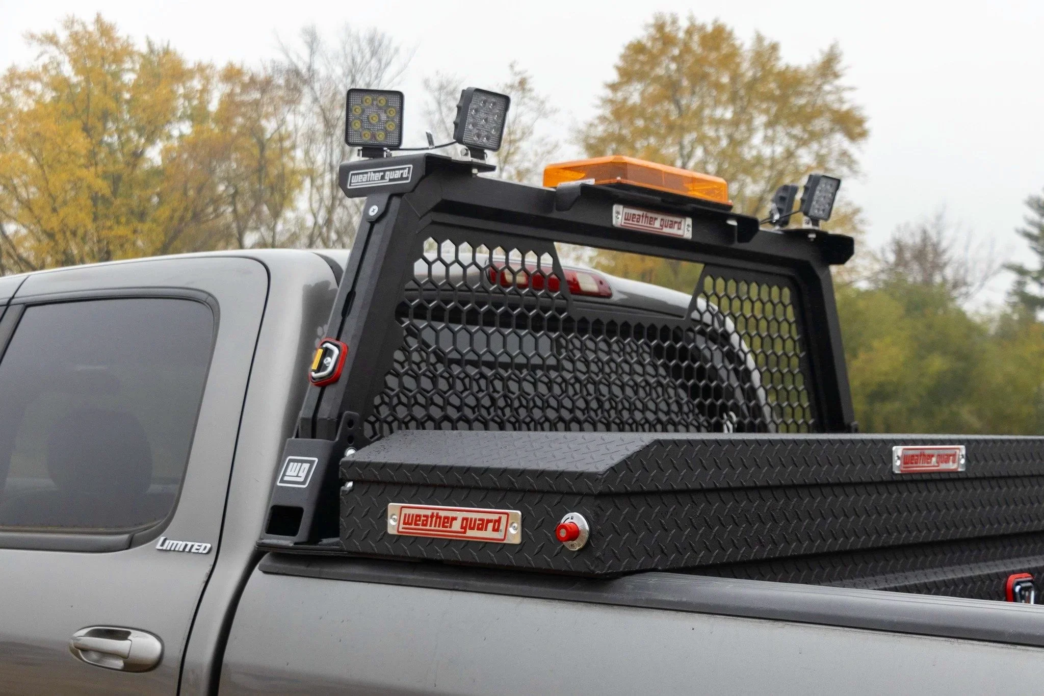 A pickup truck with a weather guard utility rack on the back, equipped with lights and a red emergency button, parked outdoors with trees in the background.