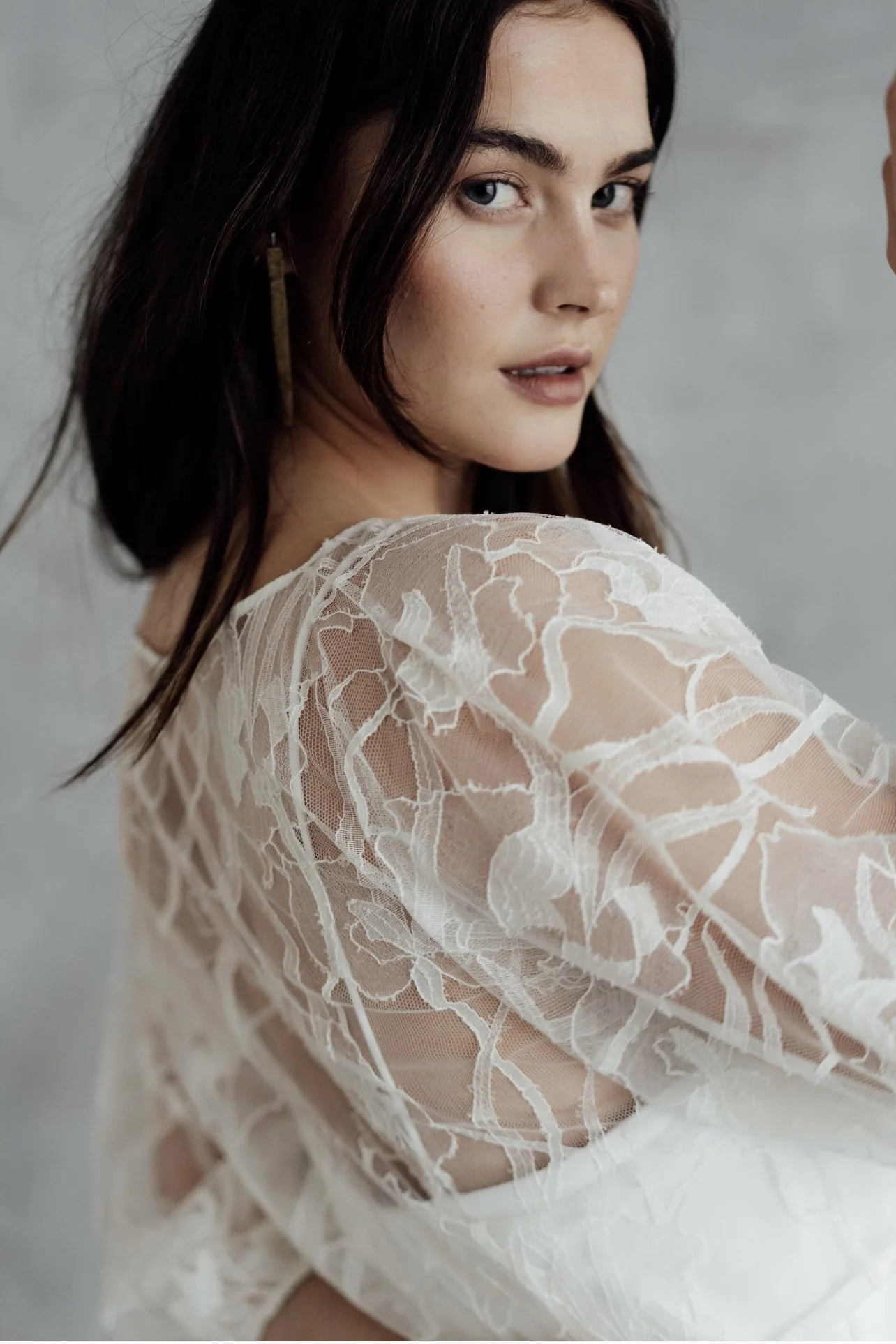 A woman with dark hair and blue eyes wearing a white lace top, looking over her shoulder at the camera.